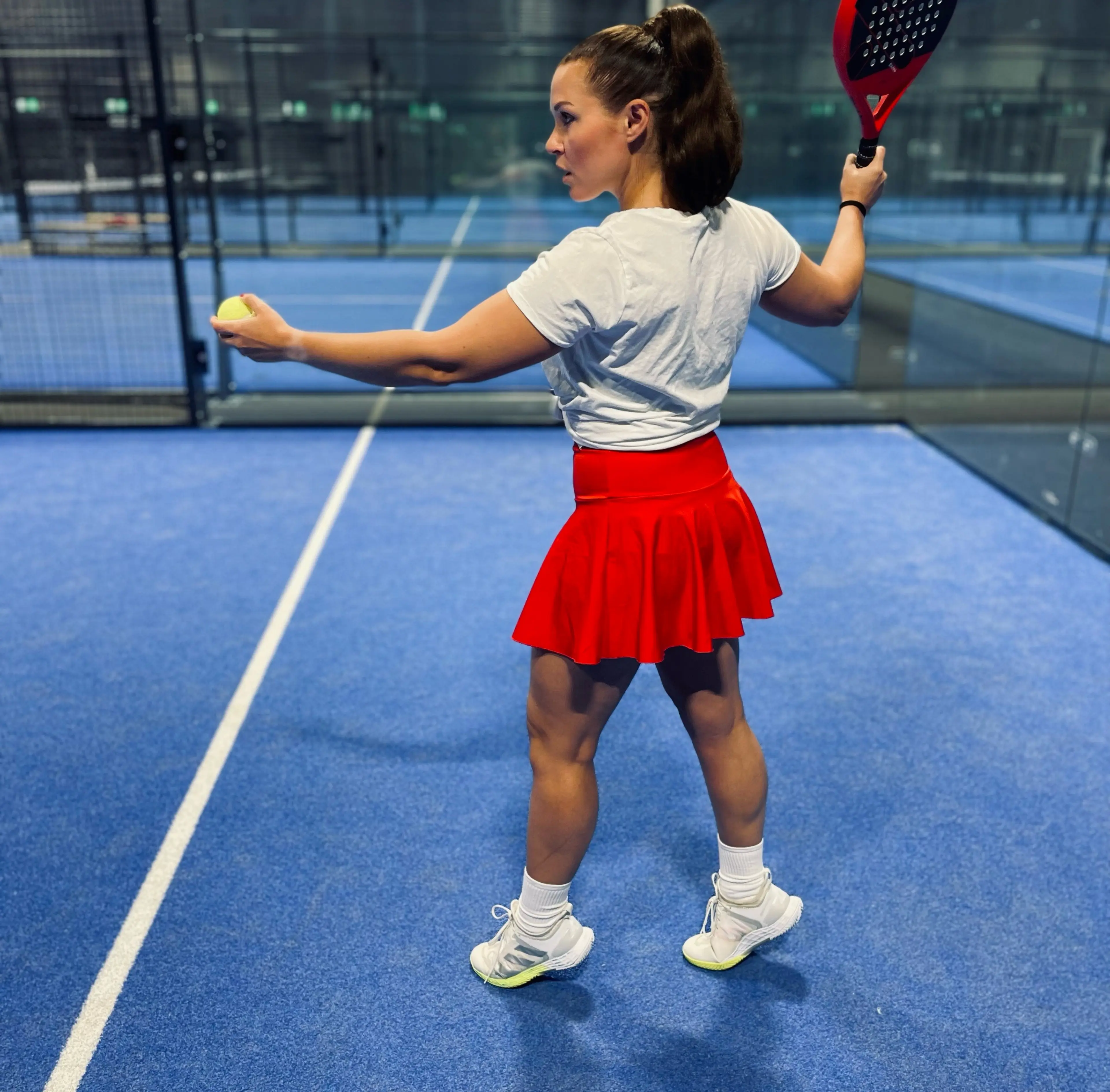 Kvinna i vit t-shirt och röd kjol håller racket redo inför serve på en blå padelbana inomhus. Hon står vänd åt sidan med en boll i handen.