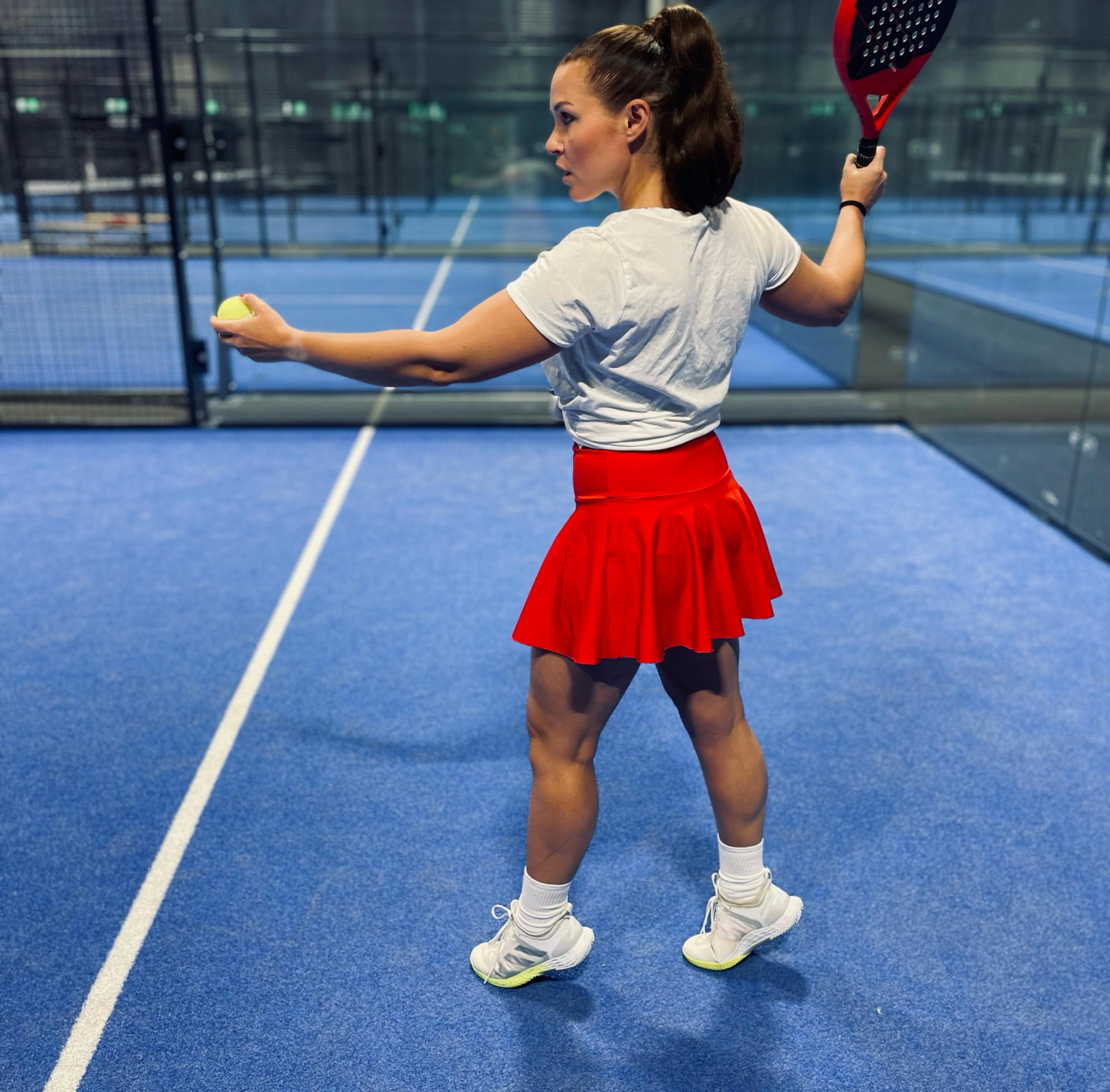 Kvinna i vit t-shirt och röd kjol håller racket redo inför serve på en blå padelbana inomhus. Hon står vänd åt sidan med en boll i handen.