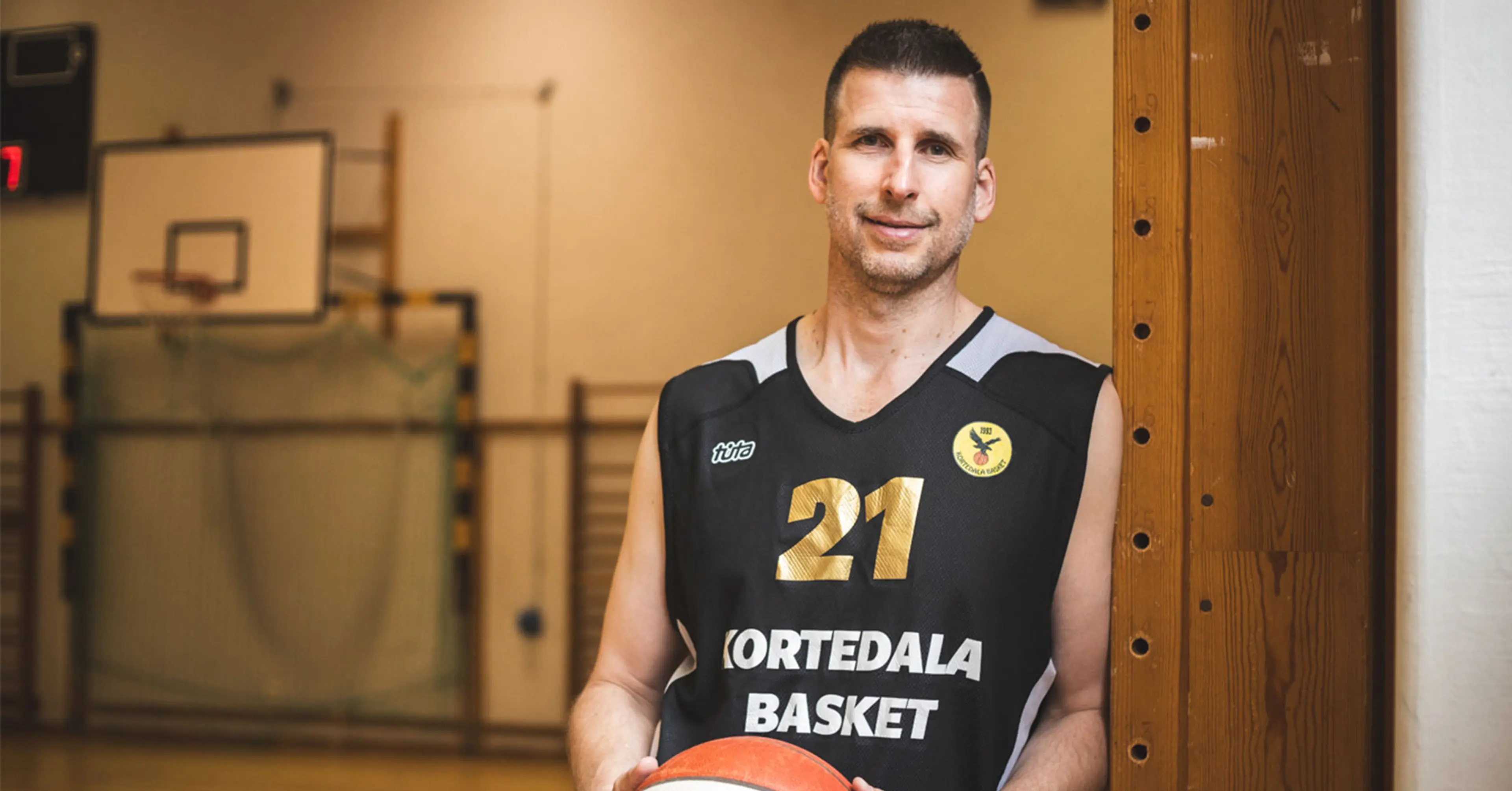 En man i svart baskettröja med nummer 21 och texten "Kortedala Basket" håller en basketboll i en sporthall. Bakgrunden visar målbur och basketkorg.