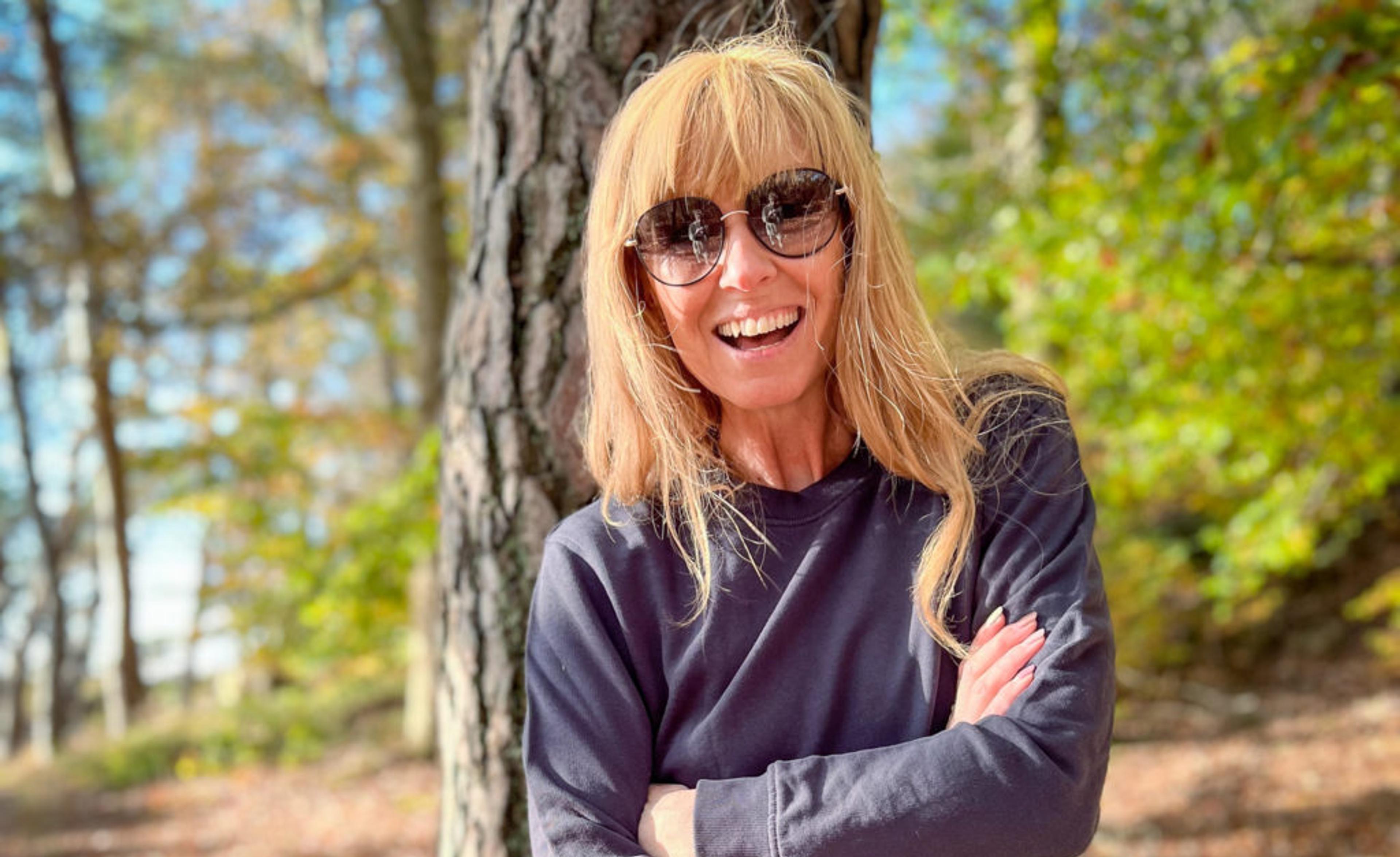 Kvinna i solglasögon och mörkgrå sweatshirt står med armarna korsade framför en trädstam i en höstskogsbacke. Ljus och skugga leker mellan träden runtomkring.
