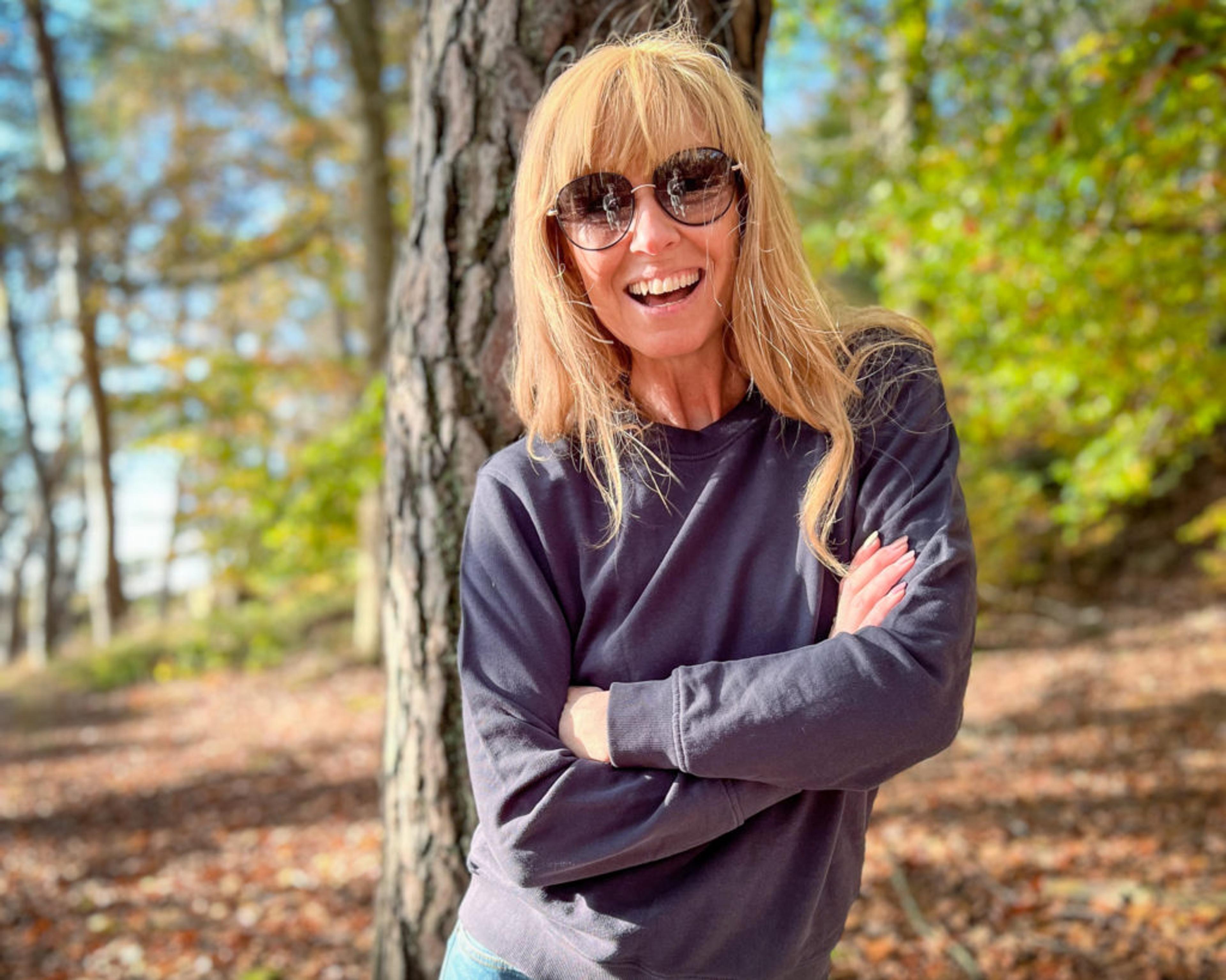 Kvinna i solglasögon och mörkgrå sweatshirt står med armarna korsade framför en trädstam i en höstskogsbacke. Ljus och skugga leker mellan träden runtomkring.