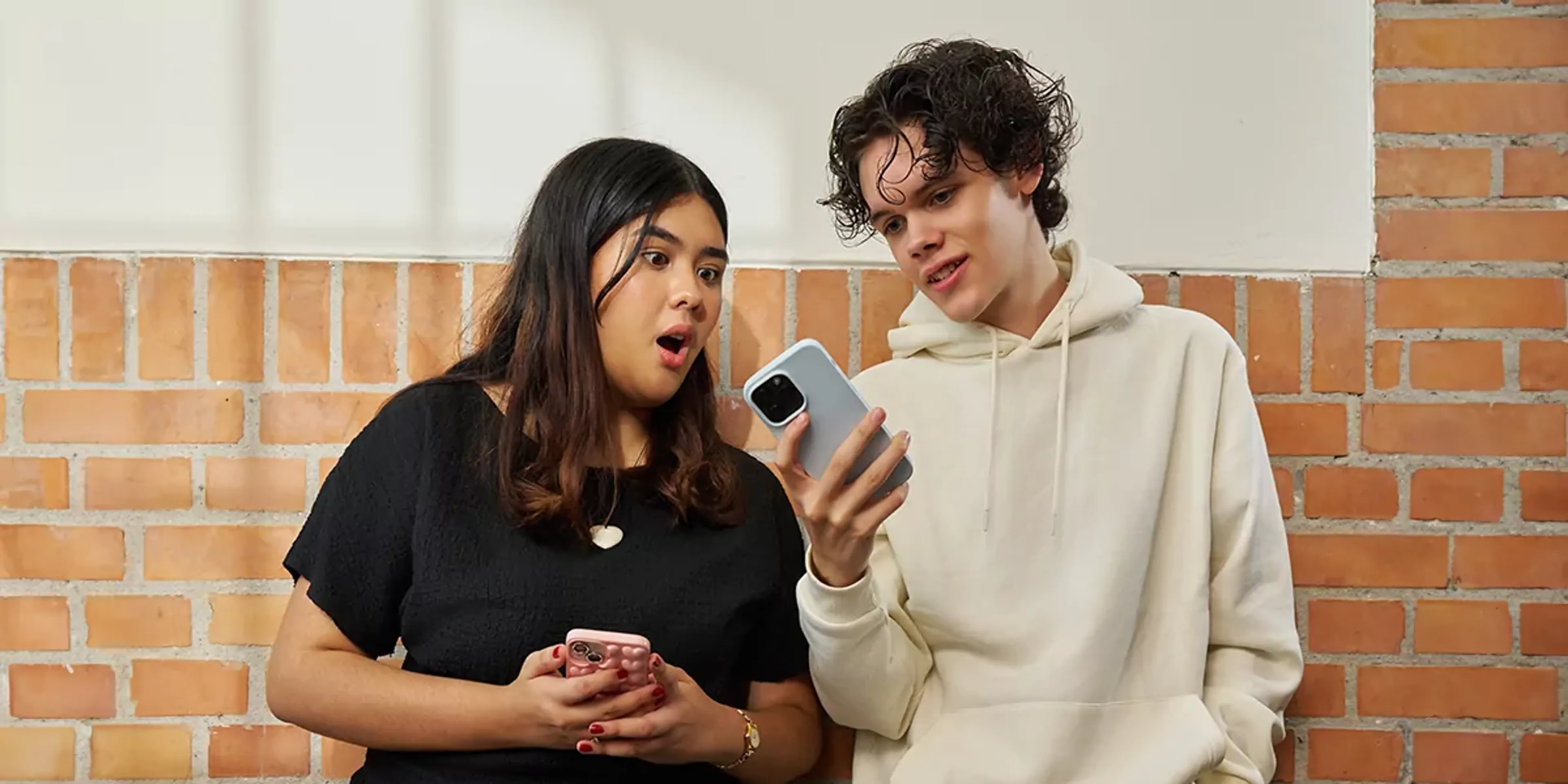 Två elever står mot en tegelvägg och tittar båda på en smartphone med intresse, den ena ser förvånad ut. De båda håller varsin mobil i händerna.