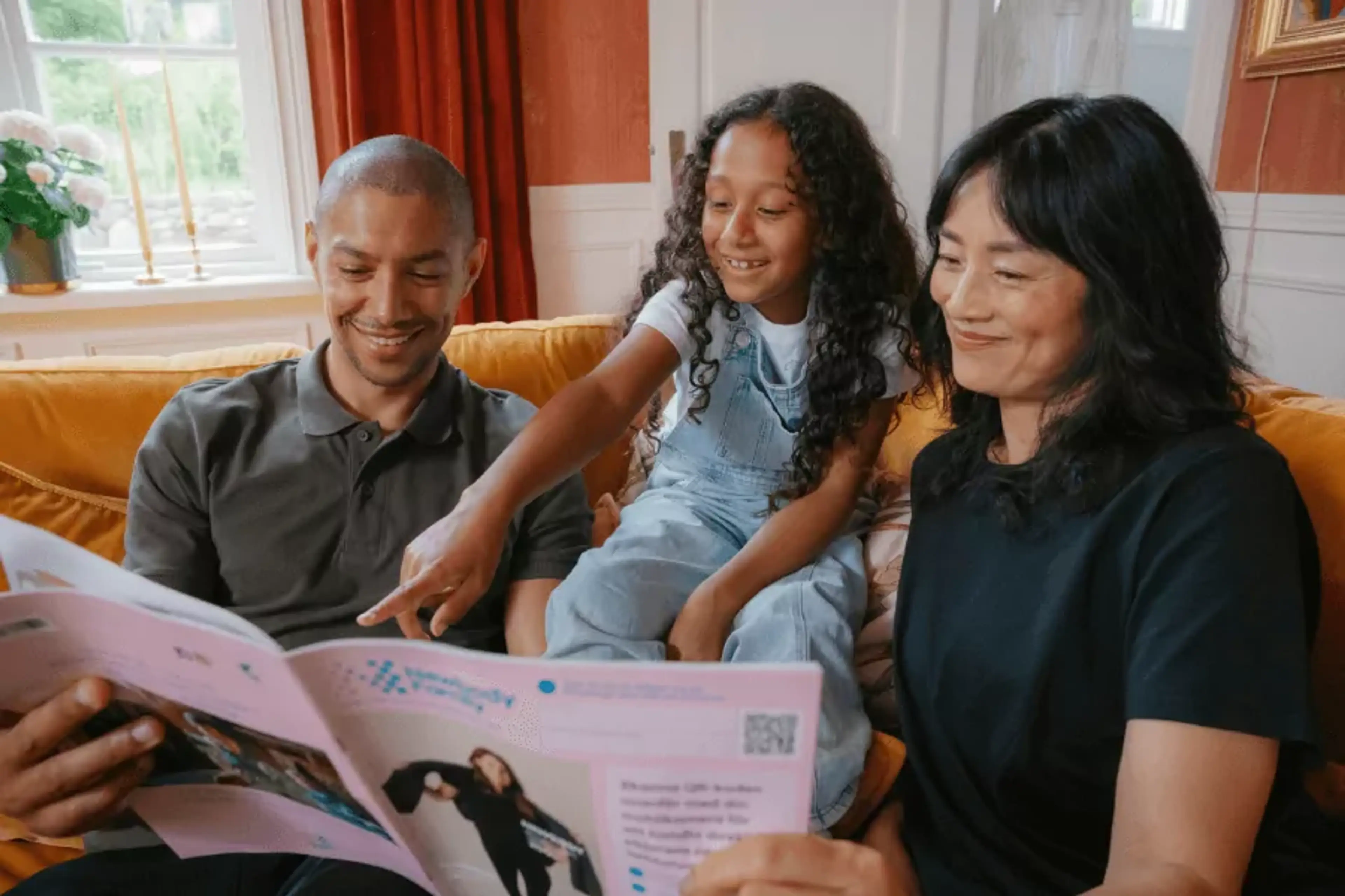 En familj sitter tillsammans i en soffa och tittar på en katalog med intresse och glädje. Barnet pekar på något i katalogen medan de vuxna ler.