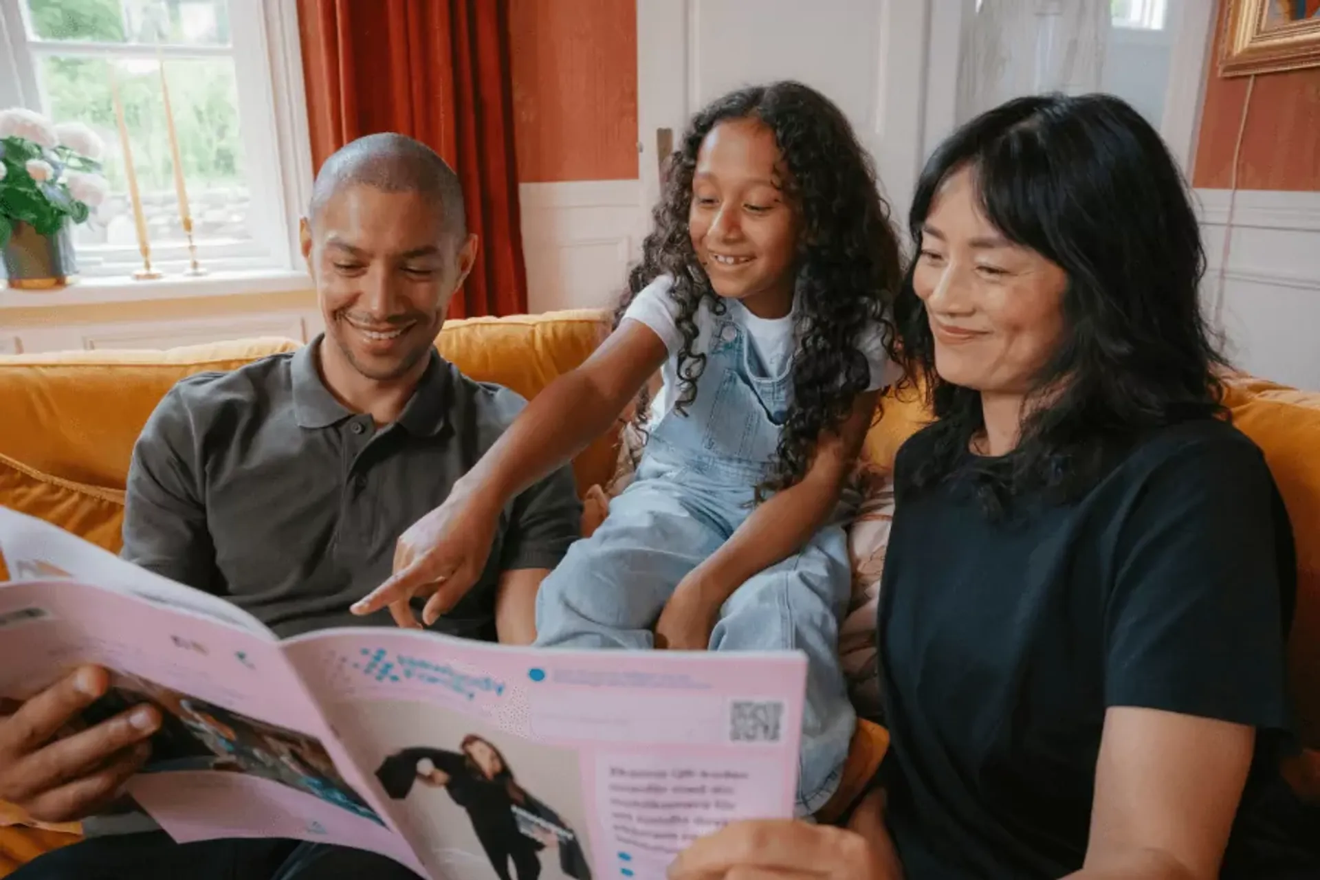 En familj sitter tillsammans i en soffa och tittar på en katalog med intresse och glädje. Barnet pekar på något i katalogen medan de vuxna ler.