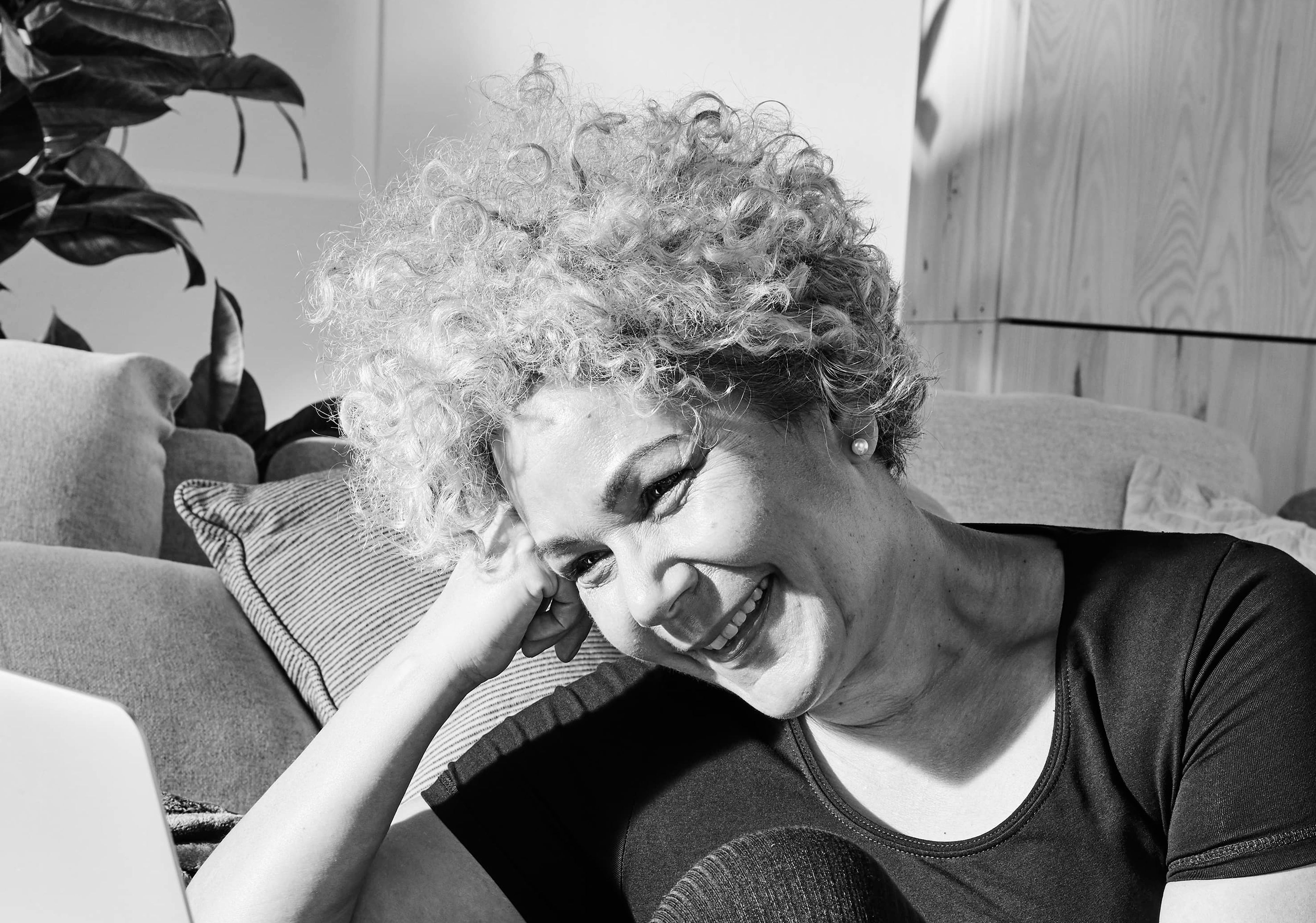 En person sitter avslappnat på golvet med böjd knä vid en soffa och tittar leende på en öppen laptop. Rummet är ombonat med kuddar och växter i bakgrunden.