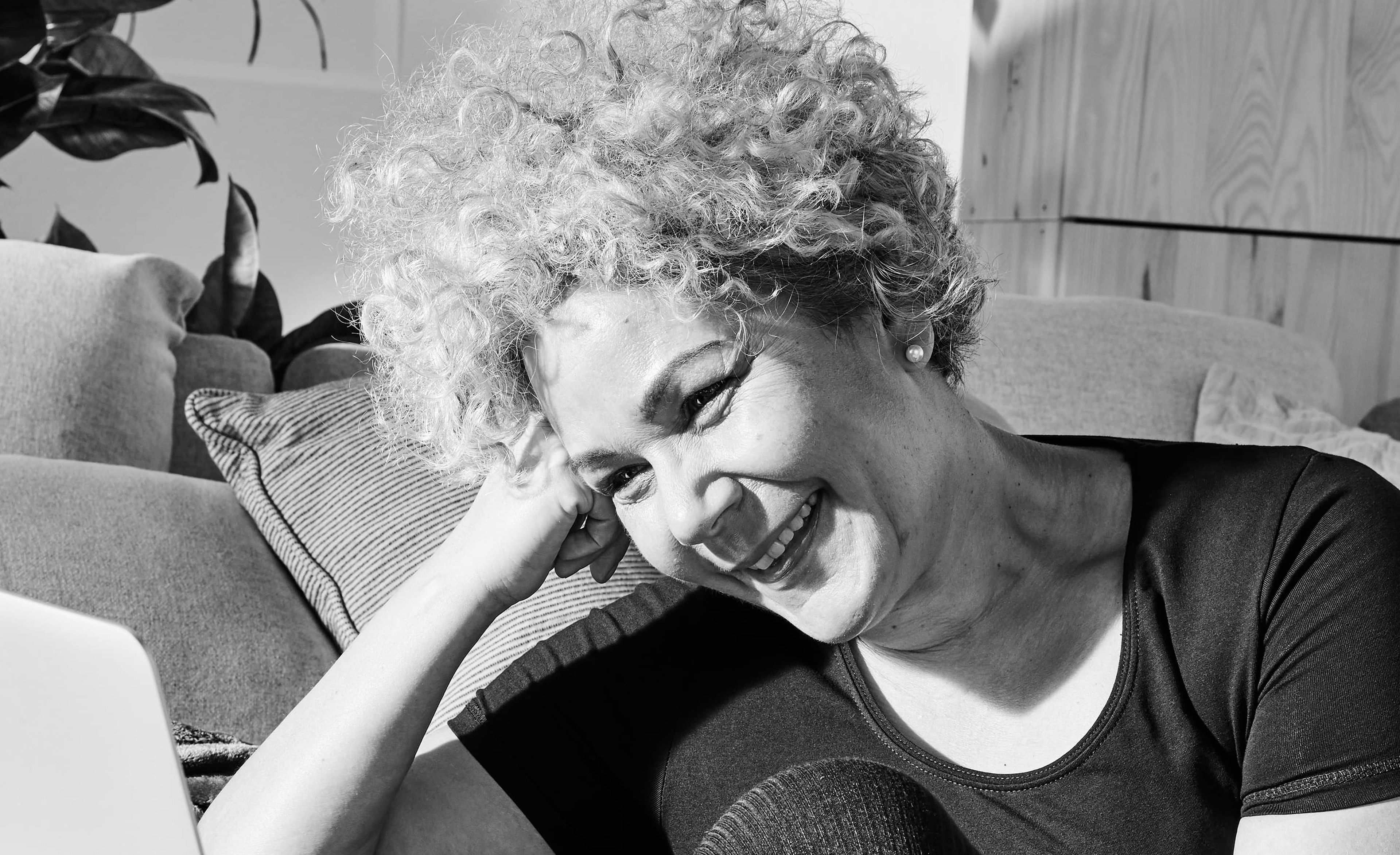 En person sitter avslappnat på golvet med böjd knä vid en soffa och tittar leende på en öppen laptop. Rummet är ombonat med kuddar och växter i bakgrunden.