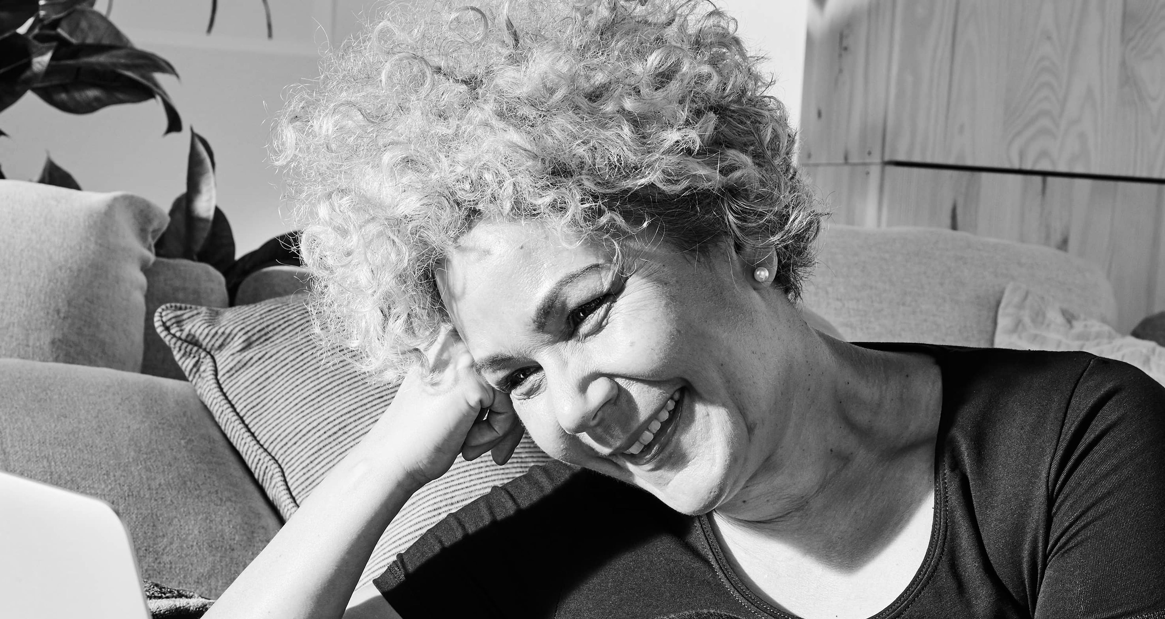 En person sitter avslappnat på golvet med böjd knä vid en soffa och tittar leende på en öppen laptop. Rummet är ombonat med kuddar och växter i bakgrunden.