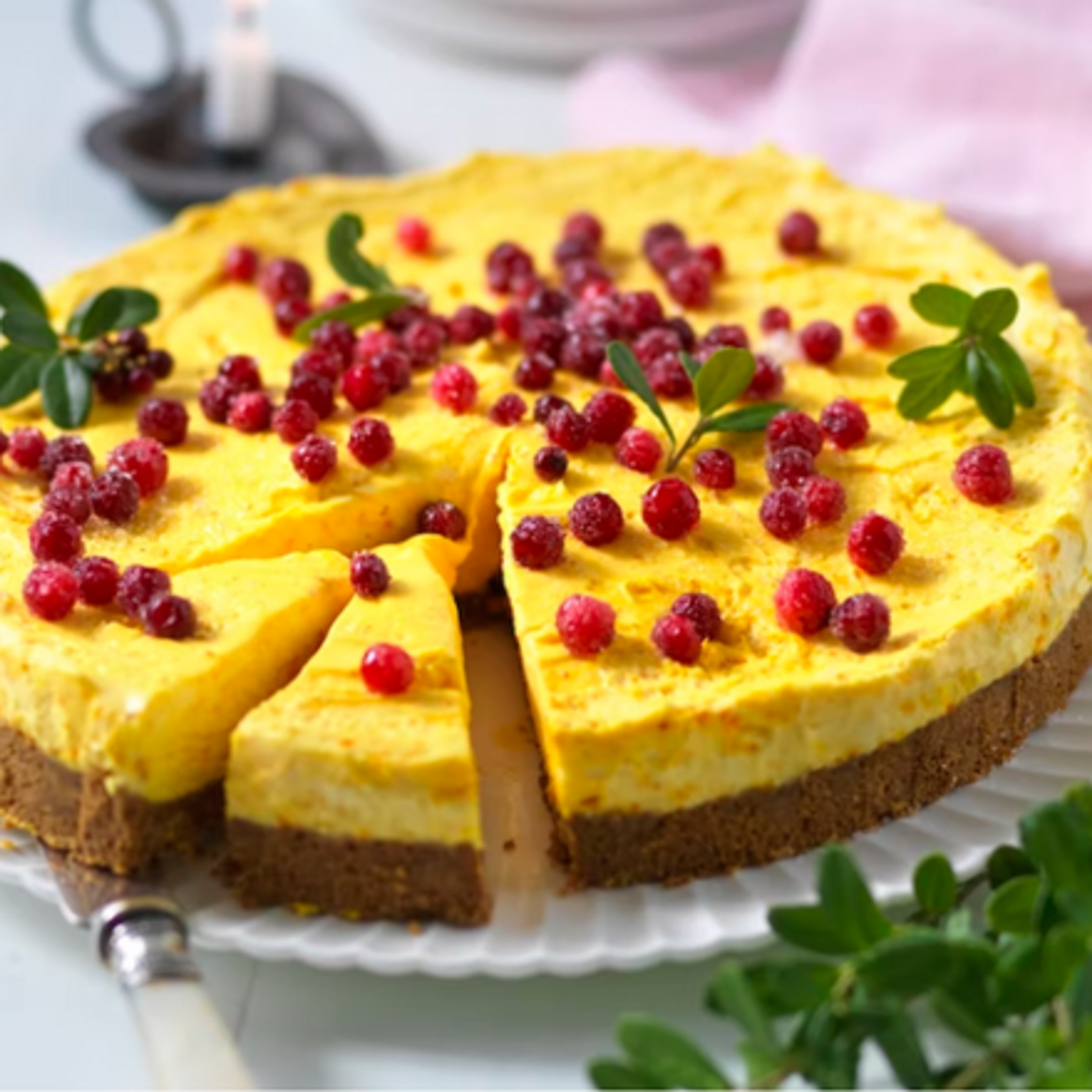En rund cheesecake med gul fyllning och krispig chokladbotten, garnerad med röda bär och några gröna blad. En bit är redan försiktigt utdragen från kakan.