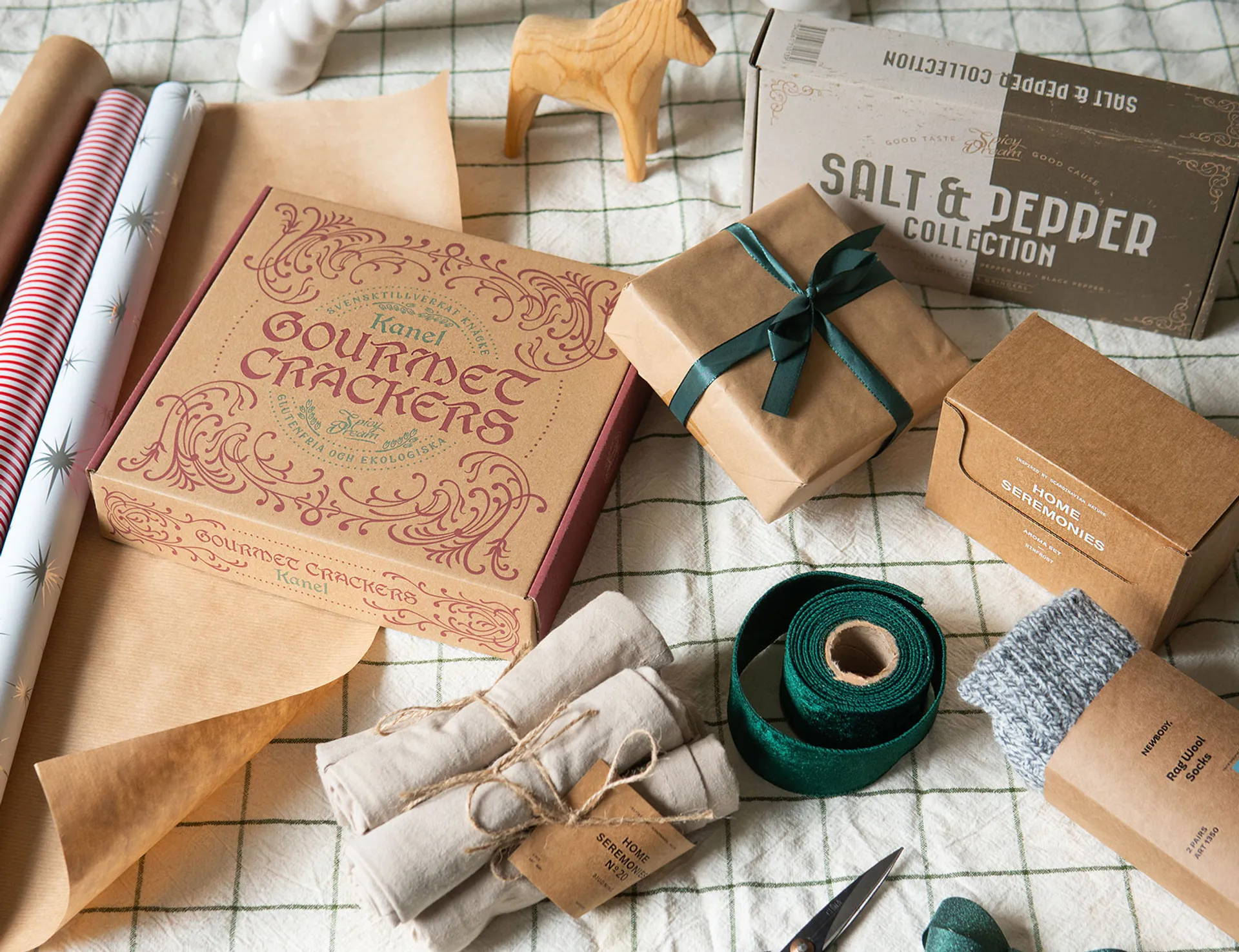 En samling presentförpackningar med gourmetkex, kryddblandningar, ullstrumpor och presentinslagningsmaterial på ett rutigt tyg. En neutral och naturlig färgskala med inslag av vinrött, grönt och beige.