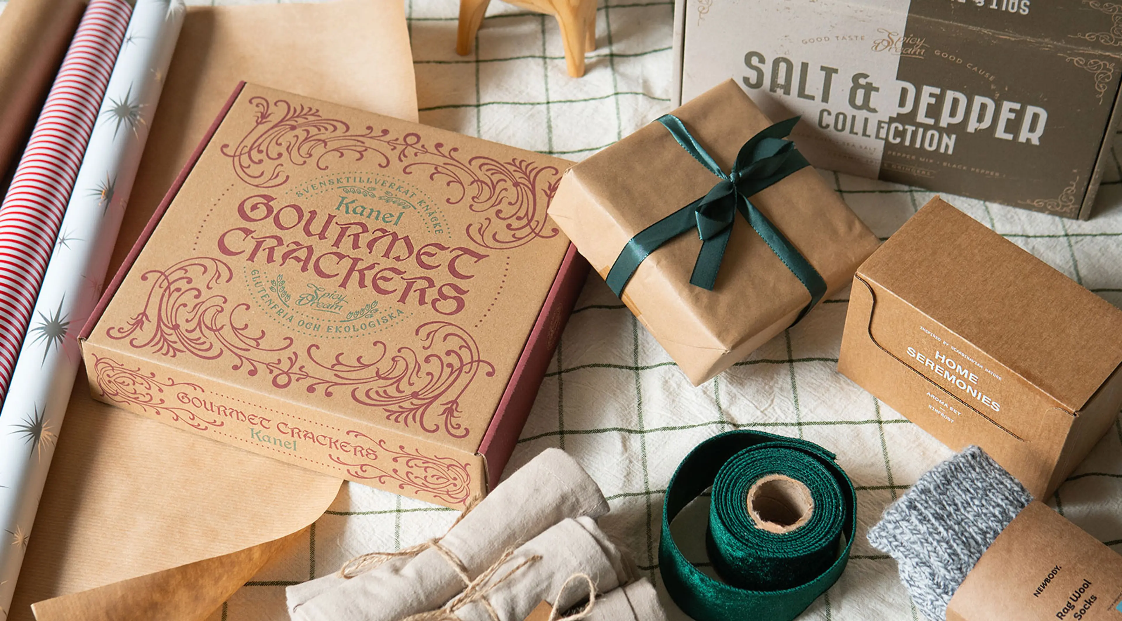 En samling presentförpackningar med gourmetkex, kryddblandningar, ullstrumpor och presentinslagningsmaterial på ett rutigt tyg. En neutral och naturlig färgskala med inslag av vinrött, grönt och beige.