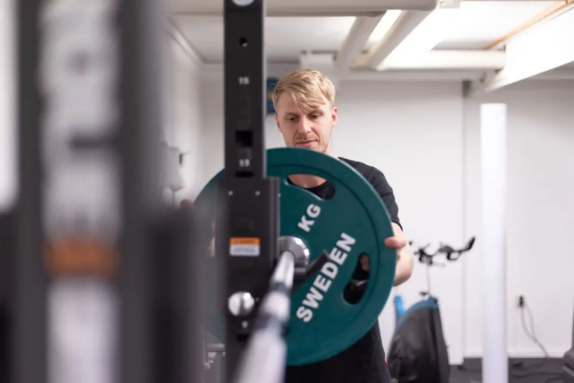 En man som sätter på en grön viktplatta på en skivstång i ett gym. I bakgrunden syns en träningscykel och vitt rum med högt i tak.