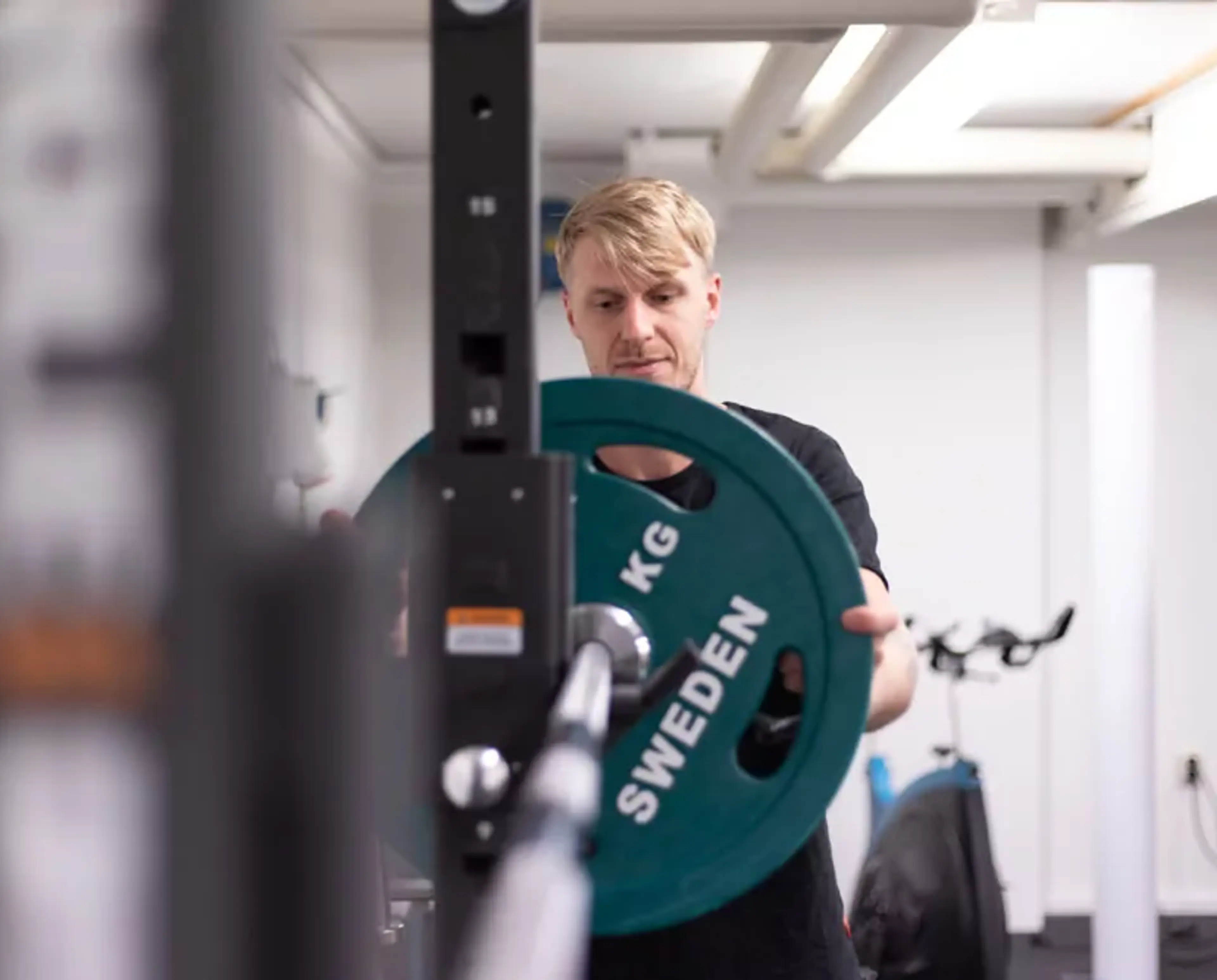 En man som sätter på en grön viktplatta på en skivstång i ett gym. I bakgrunden syns en träningscykel och vitt rum med högt i tak.