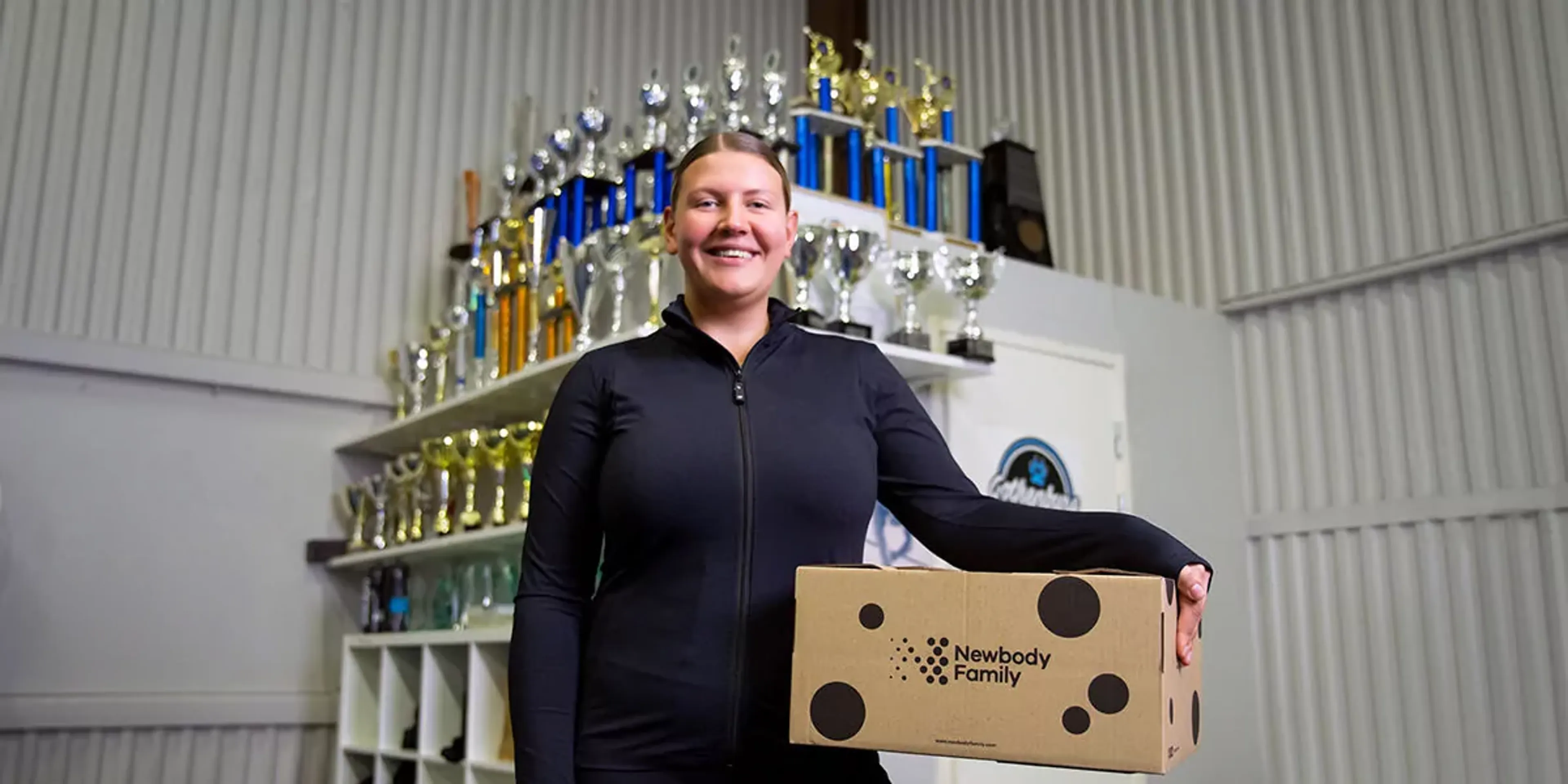 En person i svart sportjacka håller en kartong med prickar och texten "Newbody Family" i en lokal med många pokaler i bakgrunden.