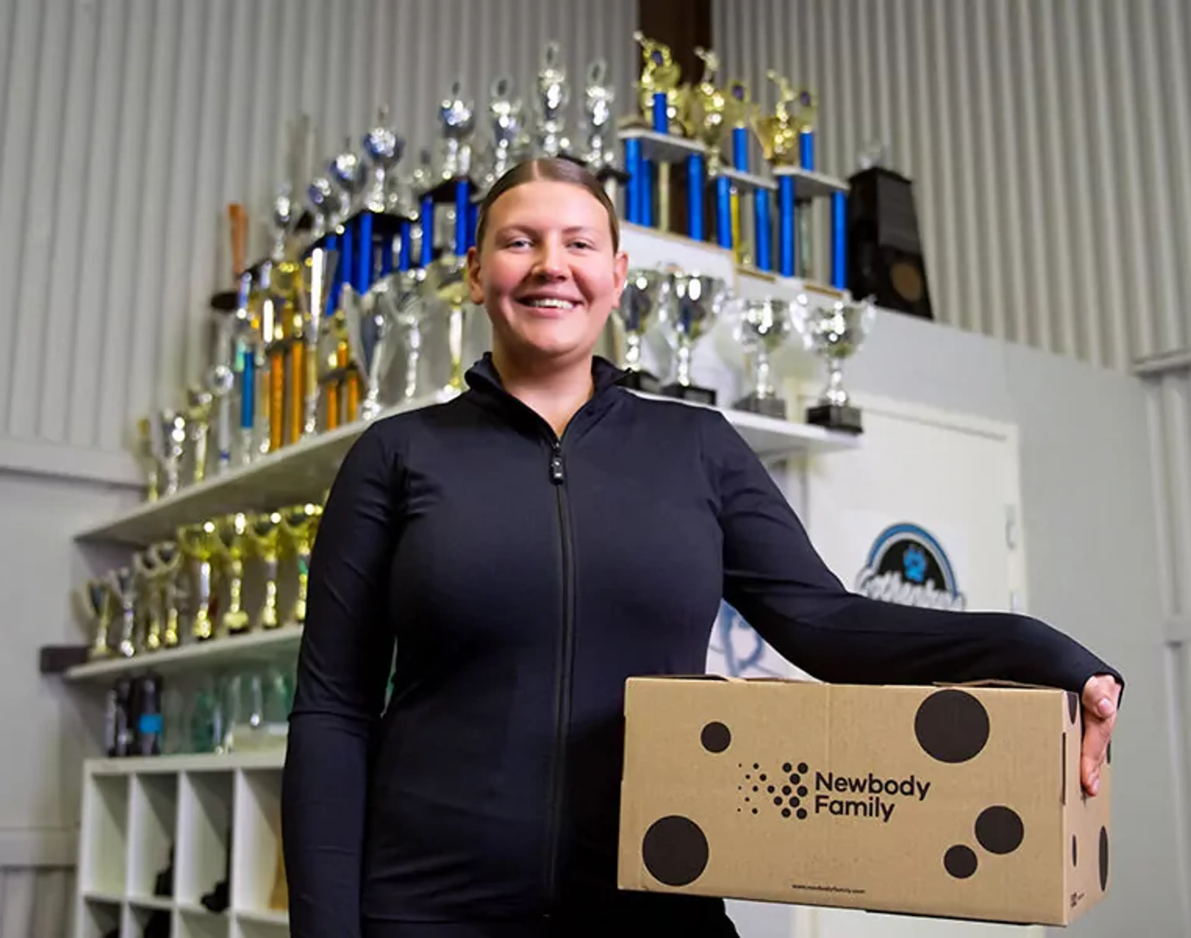 En person i svart sportjacka håller en kartong med prickar och texten "Newbody Family" i en lokal med många pokaler i bakgrunden.