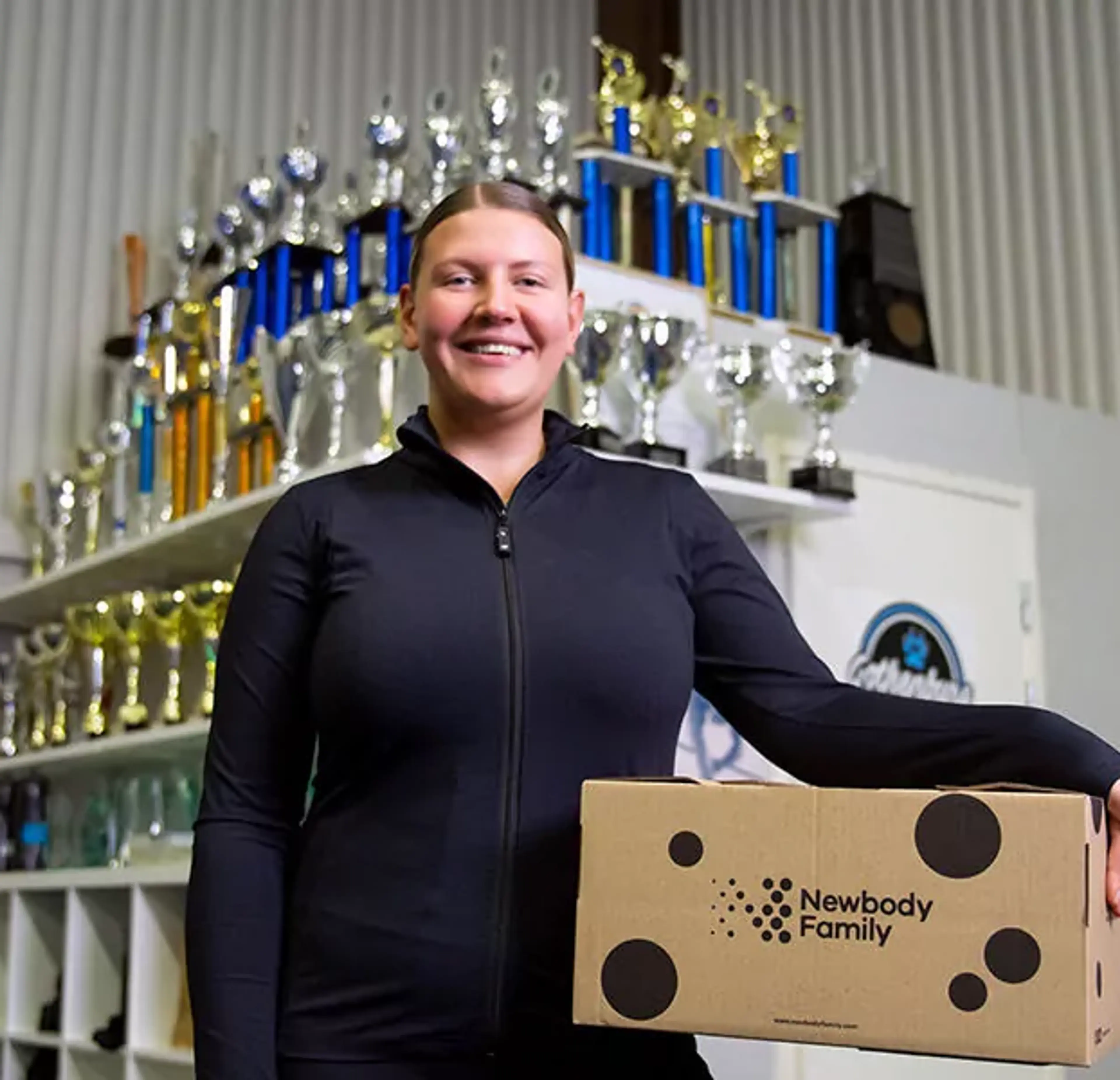 En person i svart sportjacka håller en kartong med prickar och texten "Newbody Family" i en lokal med många pokaler i bakgrunden.