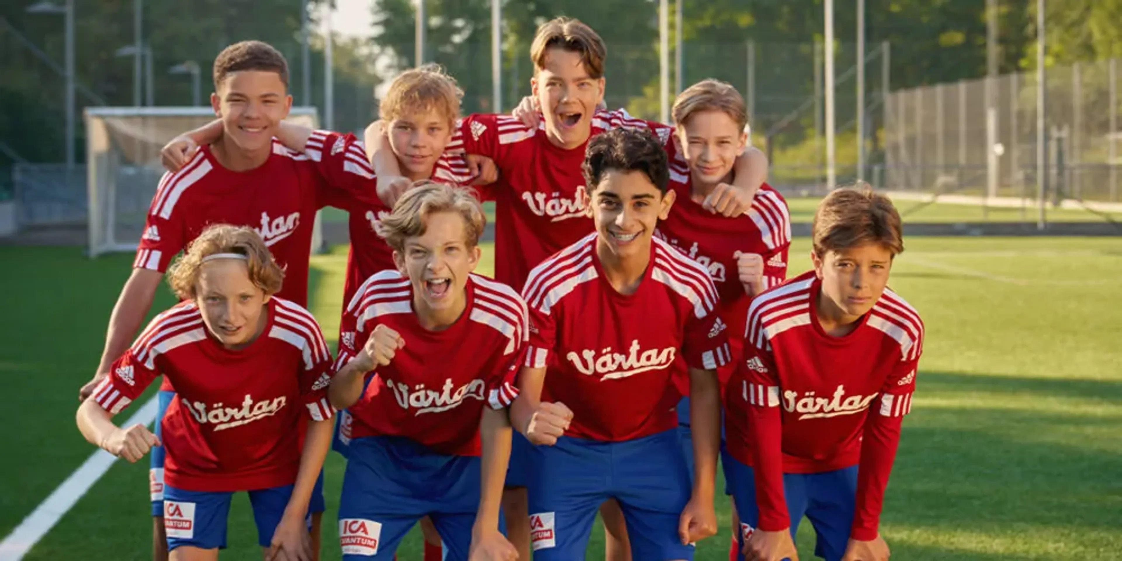 Åtta pojkar i matchkläder med röda tröjor och blå shorts står tillsammans på en fotbollsplan. De ser glada och peppade ut inför spelet.