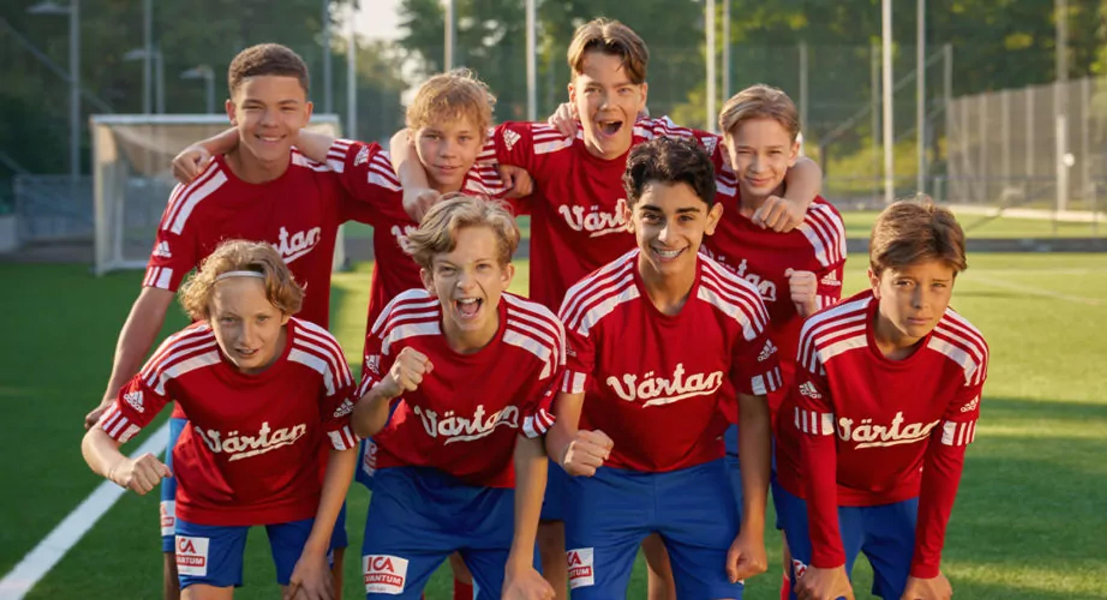 Åtta pojkar i matchkläder med röda tröjor och blå shorts står tillsammans på en fotbollsplan. De ser glada och peppade ut inför spelet.