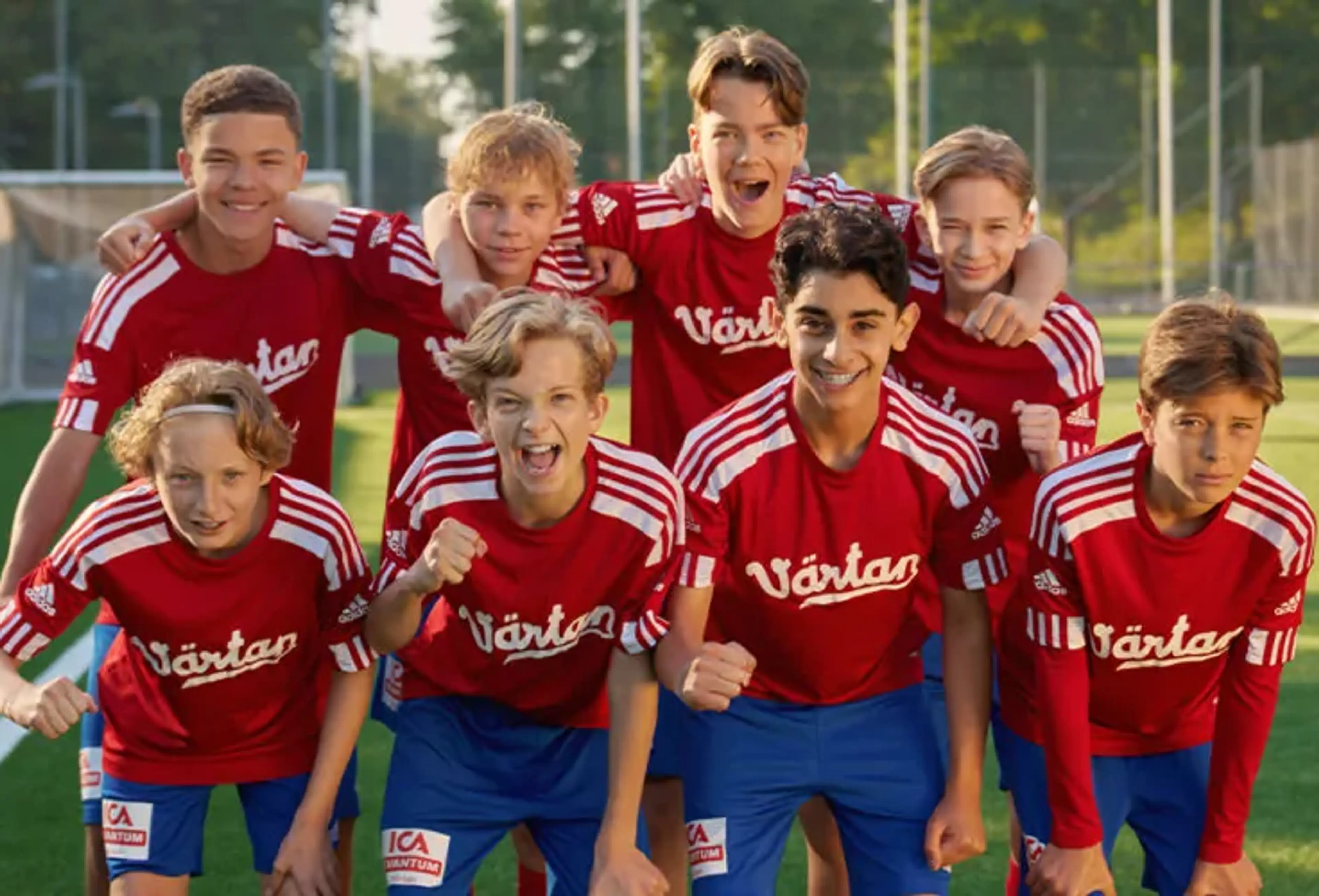 Åtta pojkar i matchkläder med röda tröjor och blå shorts står tillsammans på en fotbollsplan. De ser glada och peppade ut inför spelet.
