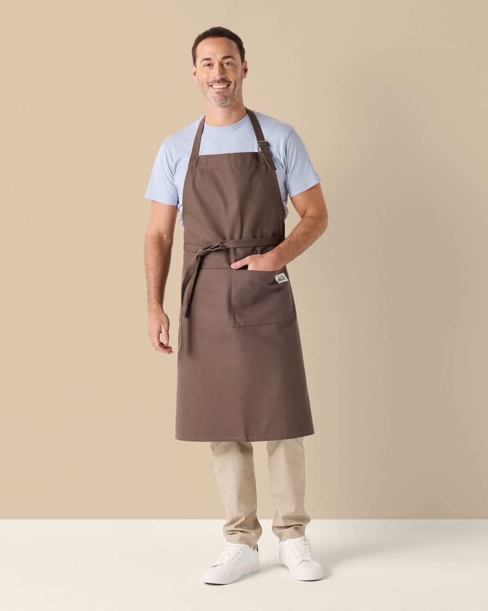 Brun förklädesmodell med justerbar nackrem och en stor framficka, bärs över ljusa byxor och en ljusblå t-shirt. Enkel och praktisk design som passar i många kök och hemmamiljöer.