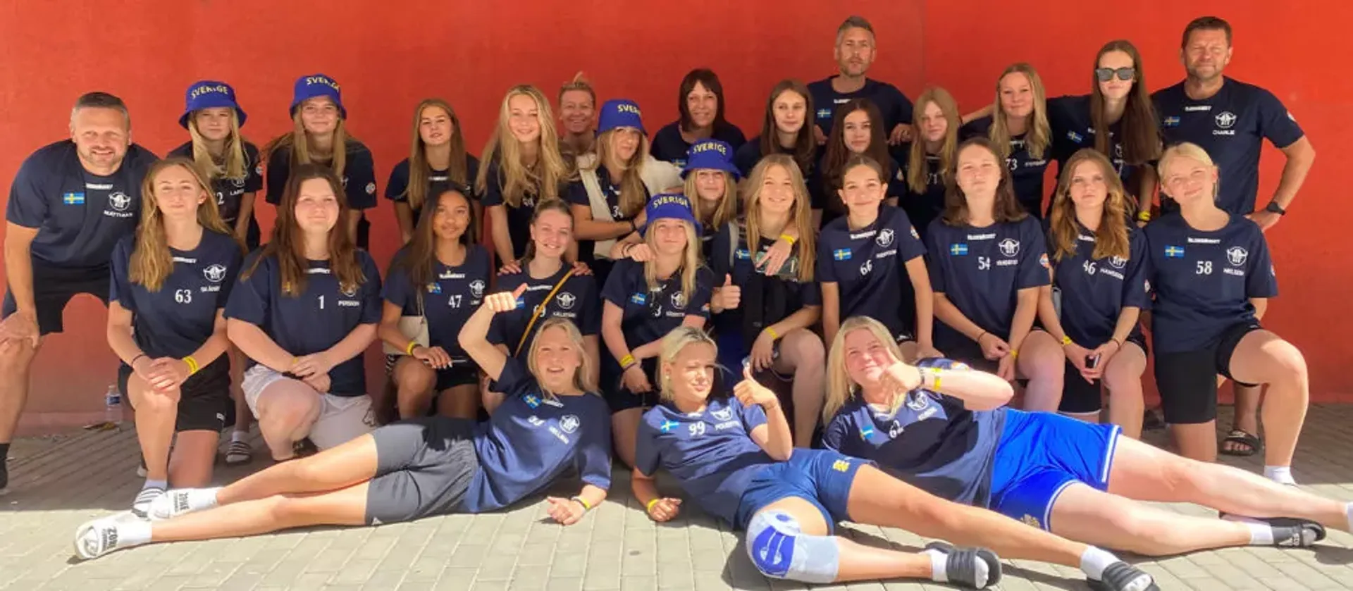 Ett stort lag med tjejer och ledare i sportkläder poserar framför en röd vägg, många med blå kepsar märkta "Sverige". Gruppen ser sammansvetsad ut och är redo för aktivitet tillsammans.