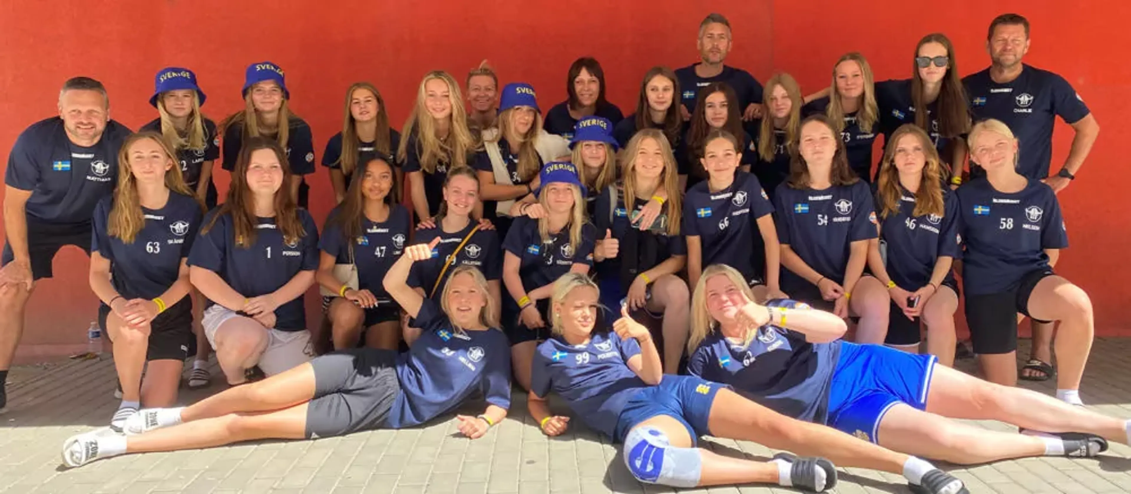 Ett stort lag med tjejer och ledare i sportkläder poserar framför en röd vägg, många med blå kepsar märkta "Sverige". Gruppen ser sammansvetsad ut och är redo för aktivitet tillsammans.