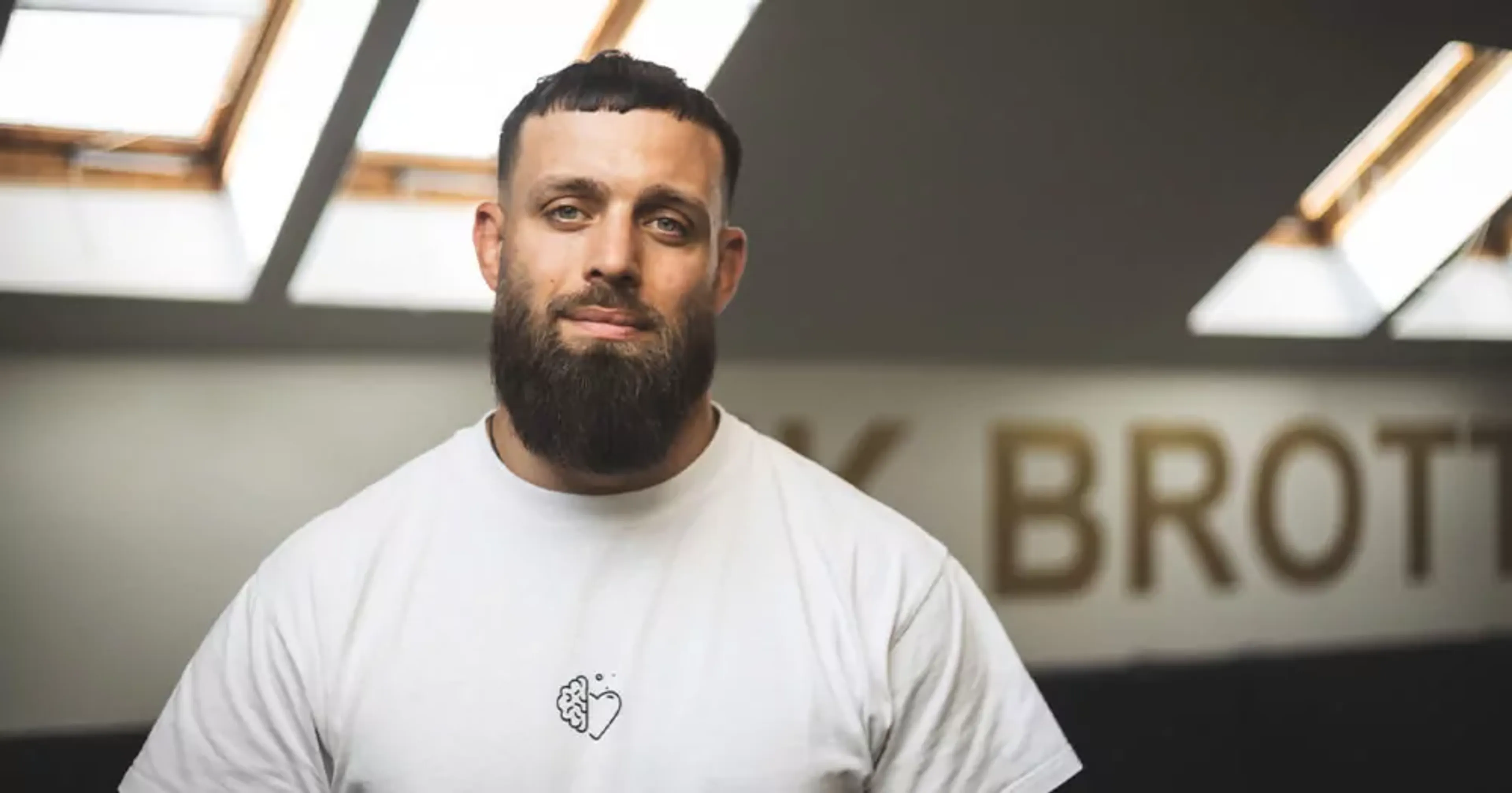 En man med kortklippt mörkt hår och skägg bär en vit t-shirt med en liten symbol föreställande ett hjärta och en hjärna på bröstet. Han står i ett rum med ljusinsläpp från takfönster och en