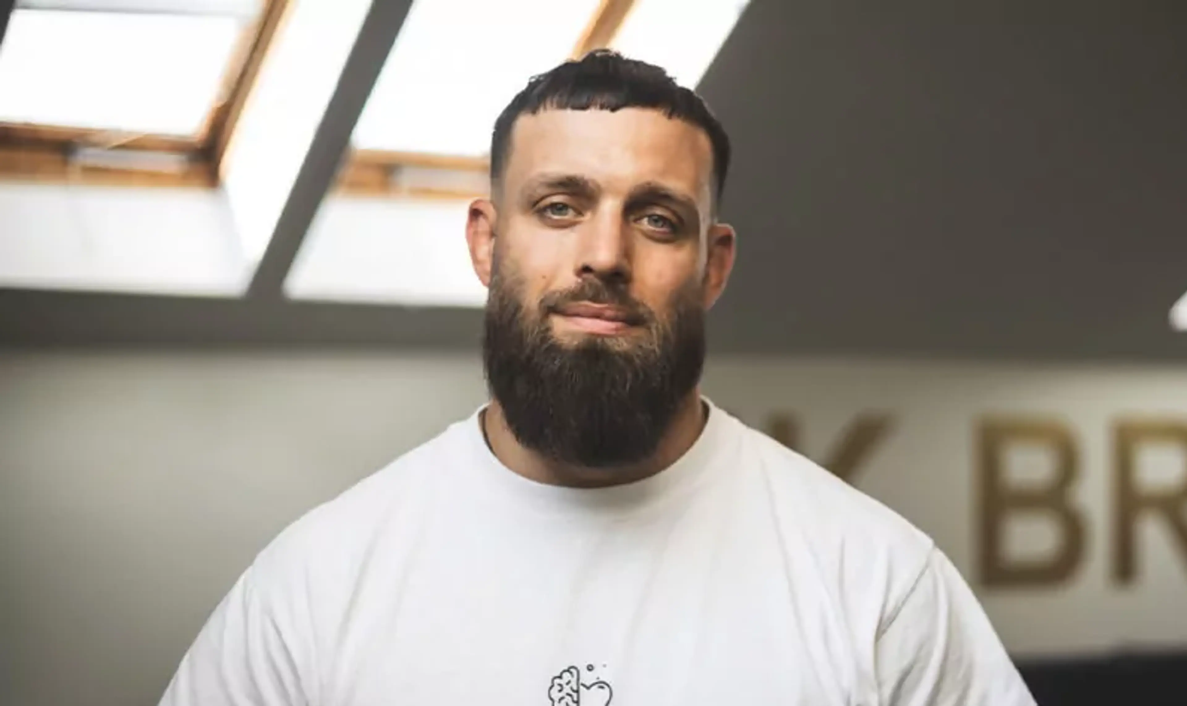 En man med kortklippt mörkt hår och skägg bär en vit t-shirt med en liten symbol föreställande ett hjärta och en hjärna på bröstet. Han står i ett rum med ljusinsläpp från takfönster och en