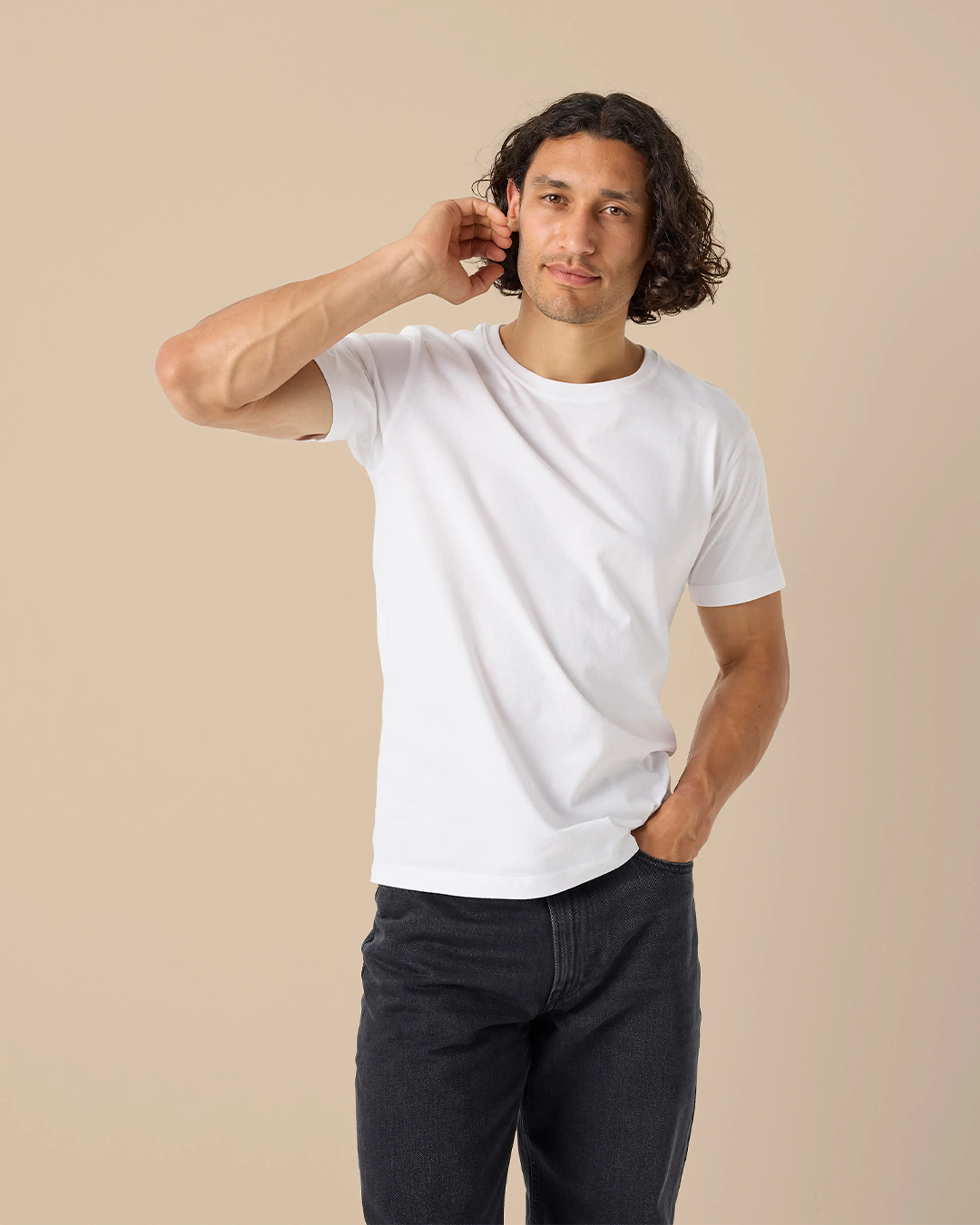 En enkel vit t-shirt i mjukt material, bärs av en person med mörkt, lockigt hår och svarta jeans. T-shirten har korta ärmar och en rund halsringning.