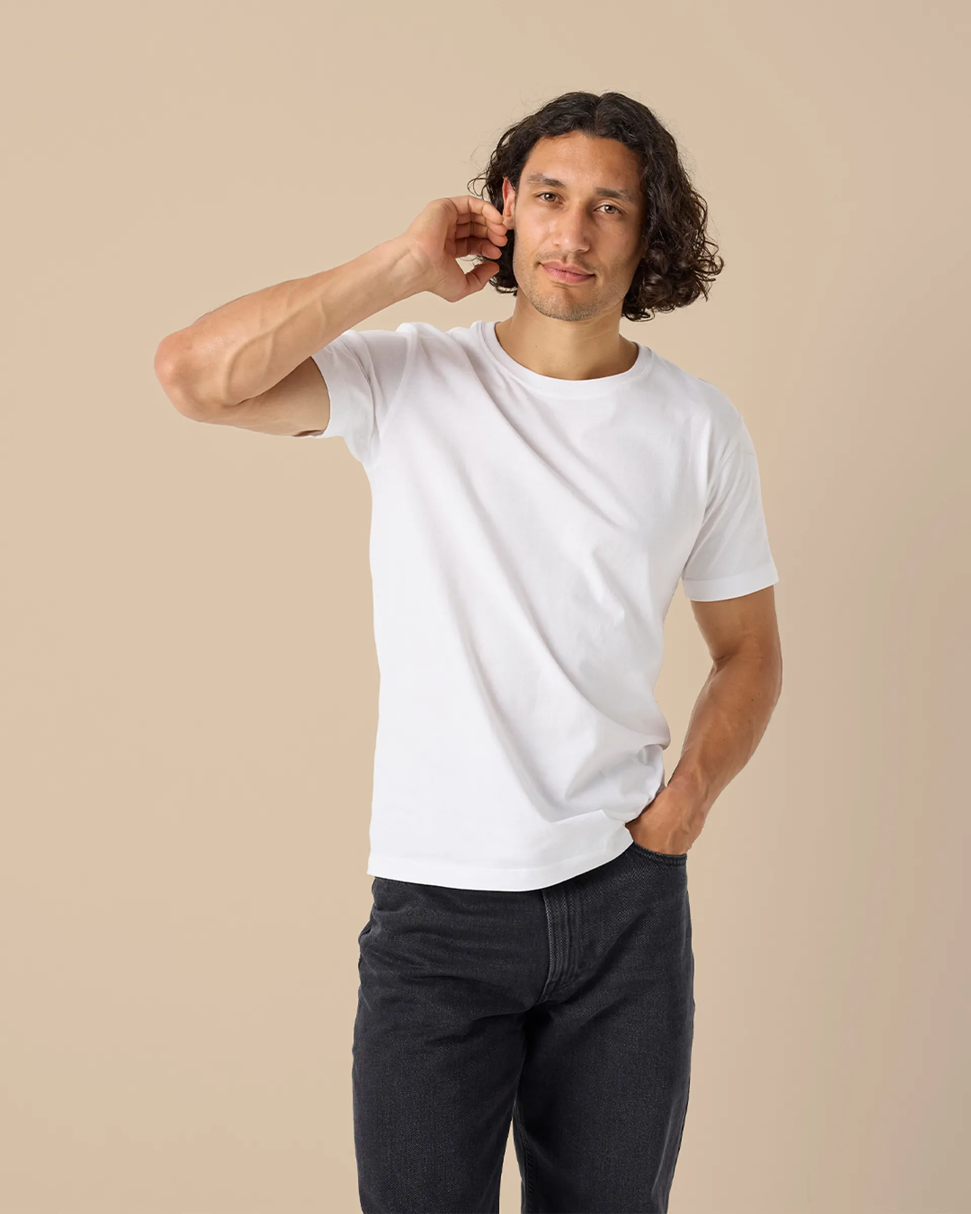 En enkel vit t-shirt i mjukt material, bärs av en person med mörkt, lockigt hår och svarta jeans. T-shirten har korta ärmar och en rund halsringning.