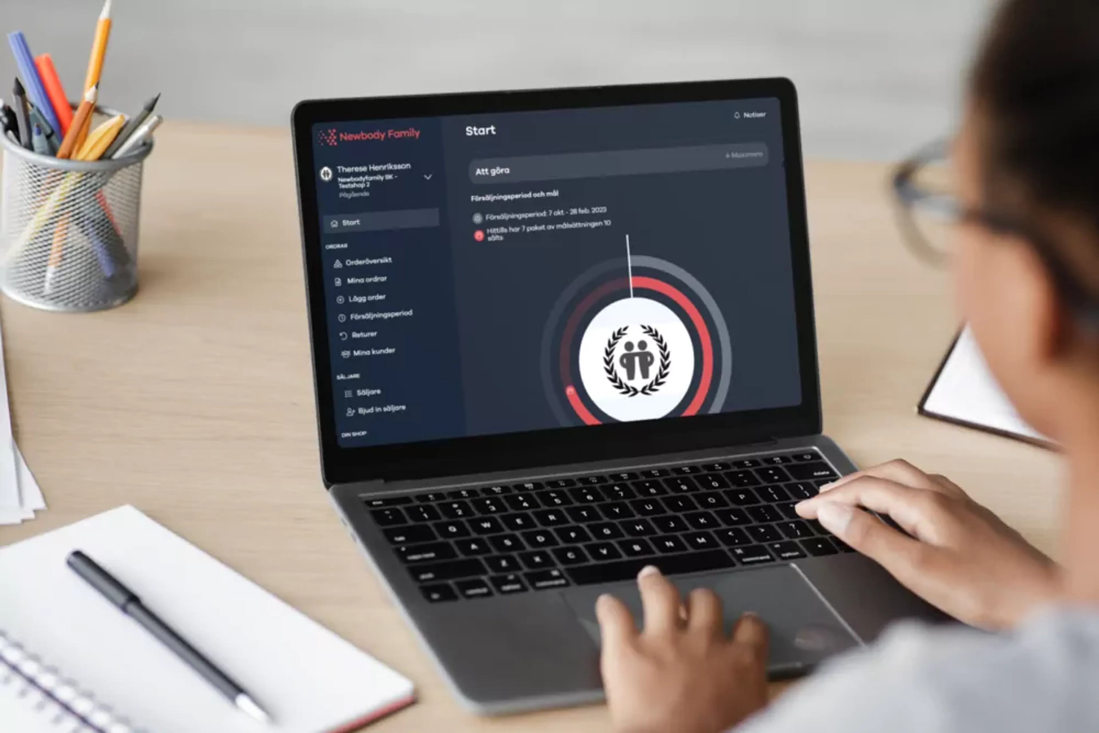 En person arbetar vid en laptop som visar en säljarplattform med uppgifter och mål. Pennor och papper ligger bredvid på skrivbordet.
