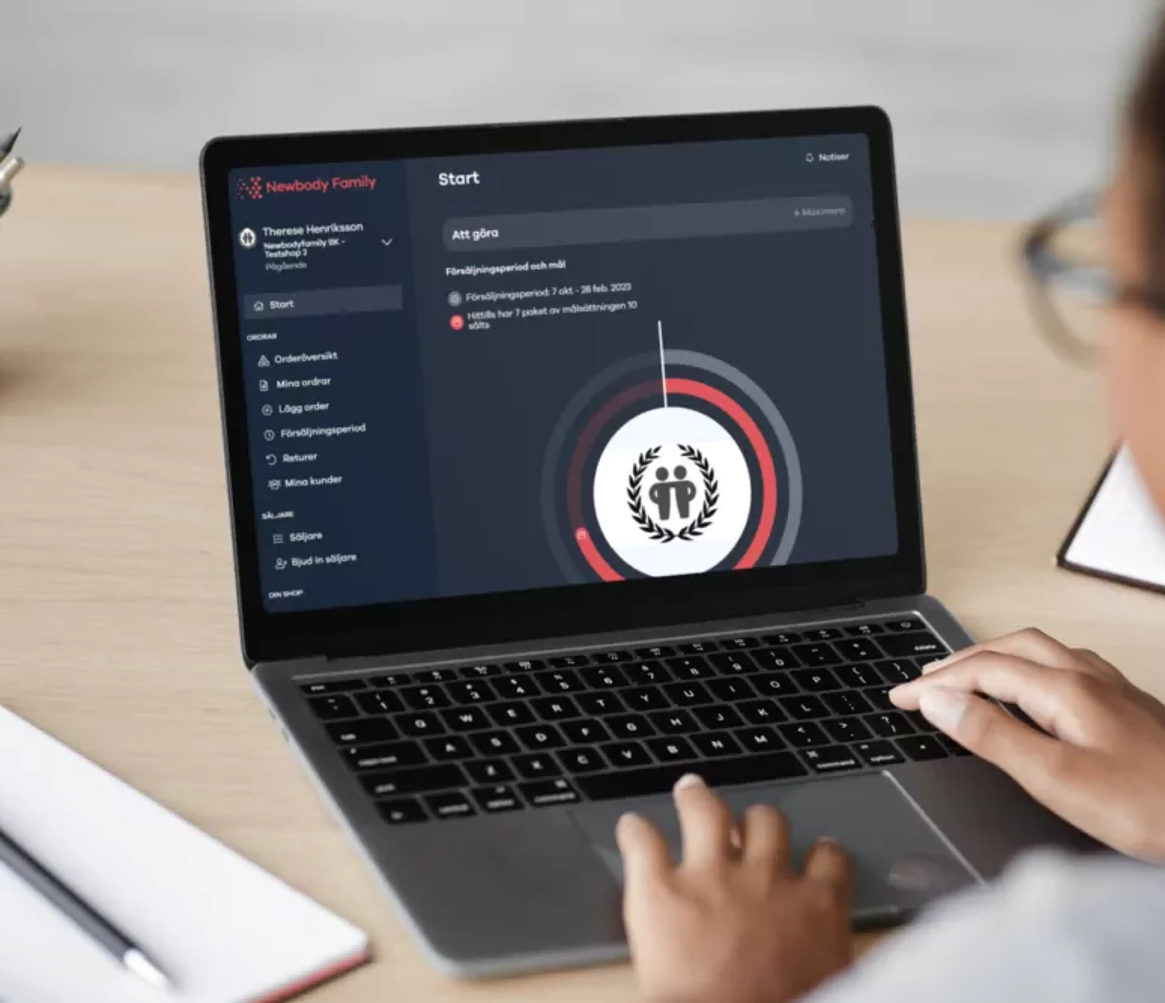 En person arbetar vid en laptop som visar en säljarplattform med uppgifter och mål. Pennor och papper ligger bredvid på skrivbordet.