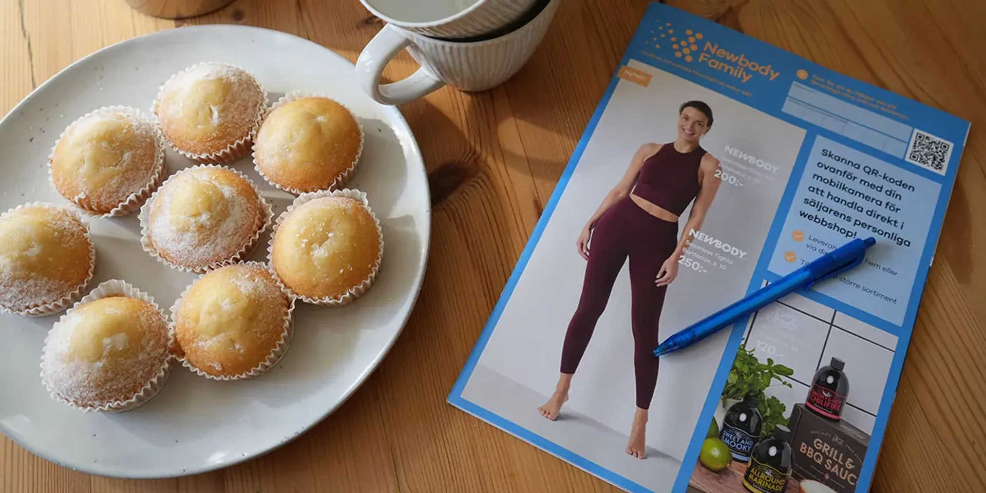 Sju små mjuka muffins med glasyr på en vit tallrik bredvid en kopp samt en katalog med träningskläder och en blå penna på ett träbord.