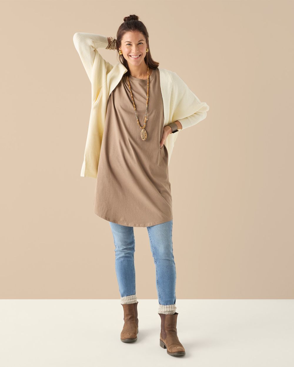 Kortärmad t-shirtklänning i mullvadsfärg på modell, stylad med jeans och stickad kofta.