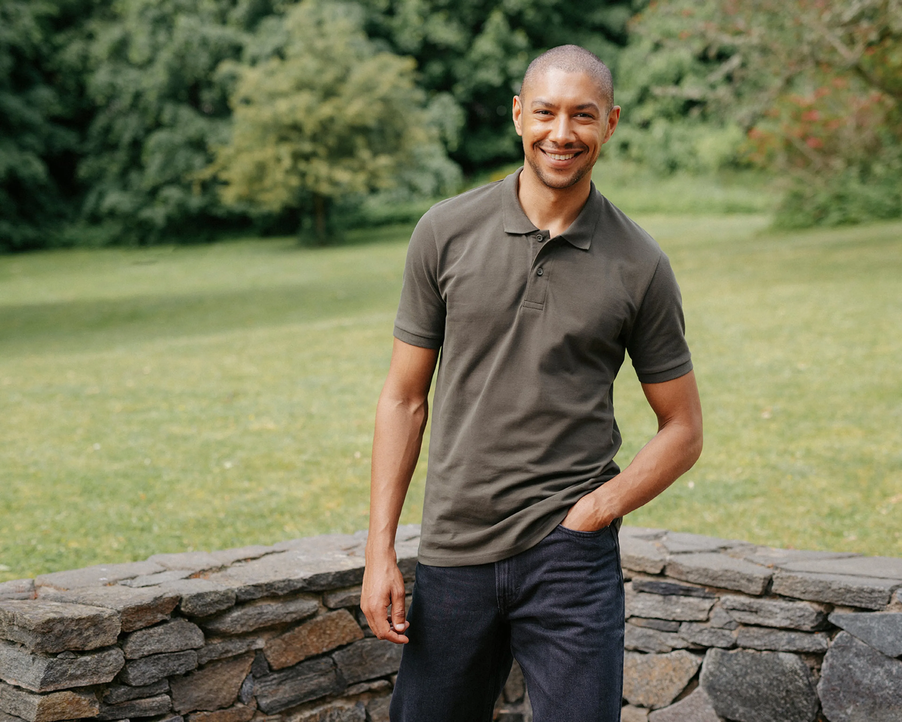 En kortärmad pikétröja i skogsgrönt med krage och knappar framtill, bärs av en leende person utomhus. Tröjan har en enkel och tidlös design.