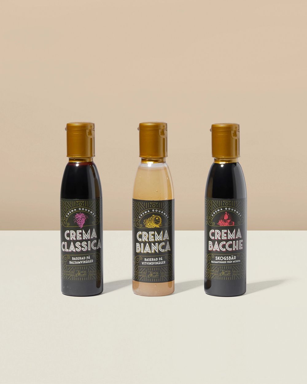 Tre små flaskor med olika balsamicokräm, en mörk klassisk, en ljus vitvinbalsam och en mörk med smak av skogsbär. Flaskorna har guldiga korkar och etiketter med dekorativa detaljer.