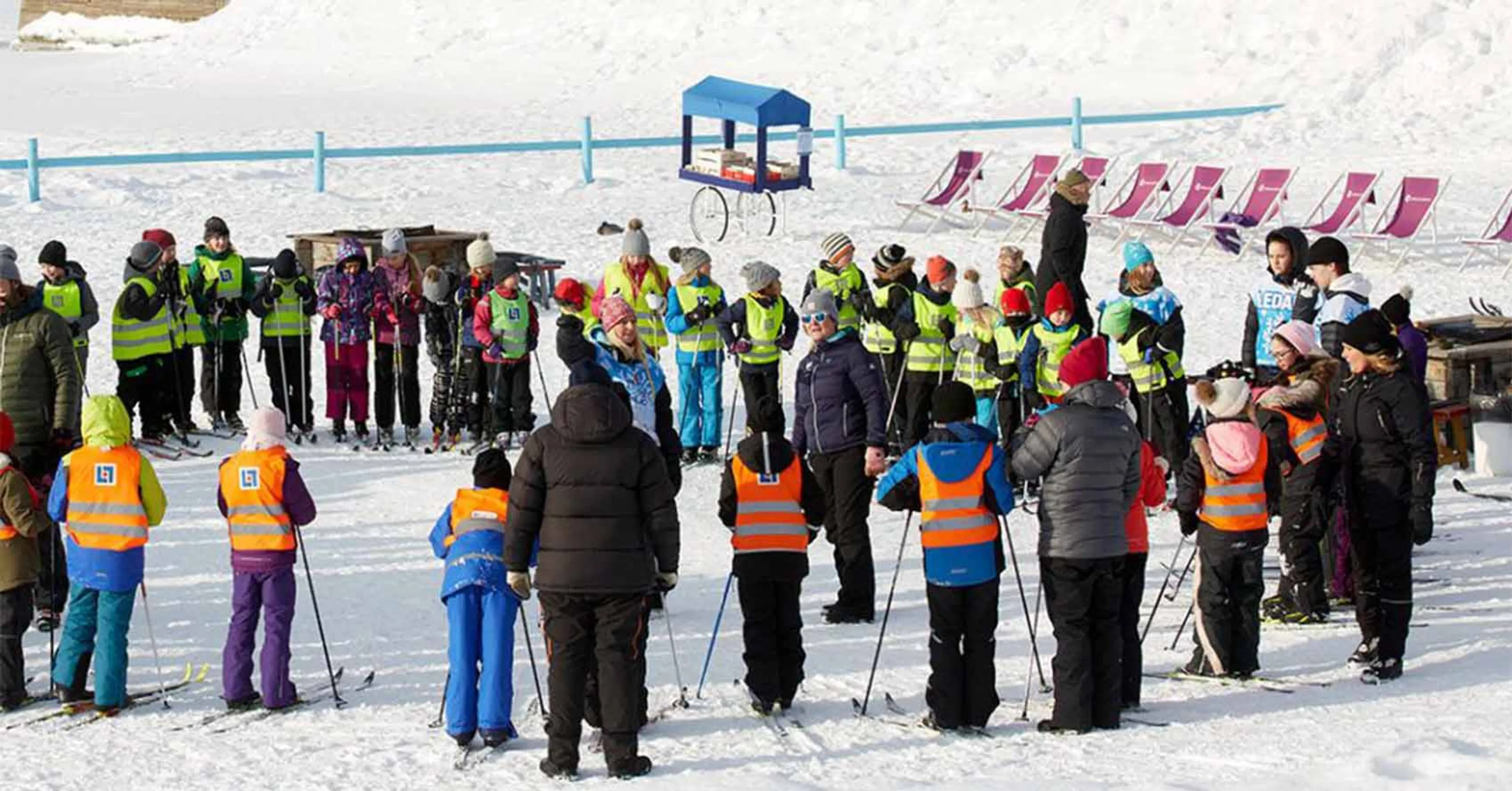 Barn och vuxna samlade i cirkel på en snötäckt yta, alla iklädda färgglada vinterkläder och säkerhetsvästar. Området är en öppen vinterscen med trädrad och byggnader i bakgrunden.