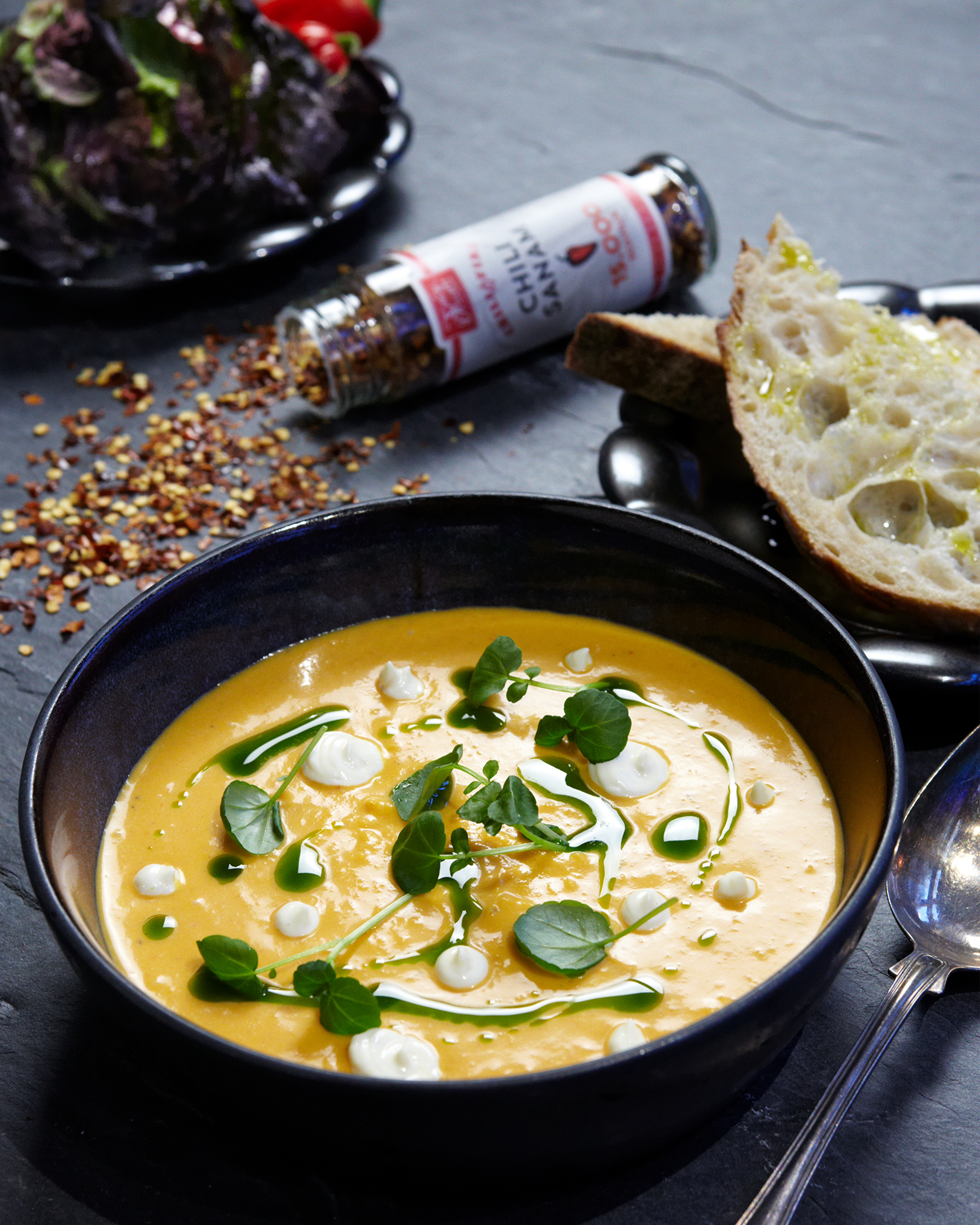 Krämig chili- och soppasoppa i mörk skål, toppad med gröna blad och klickar av grädde, med chiliflingor och två skivor bröd vid sidan om.