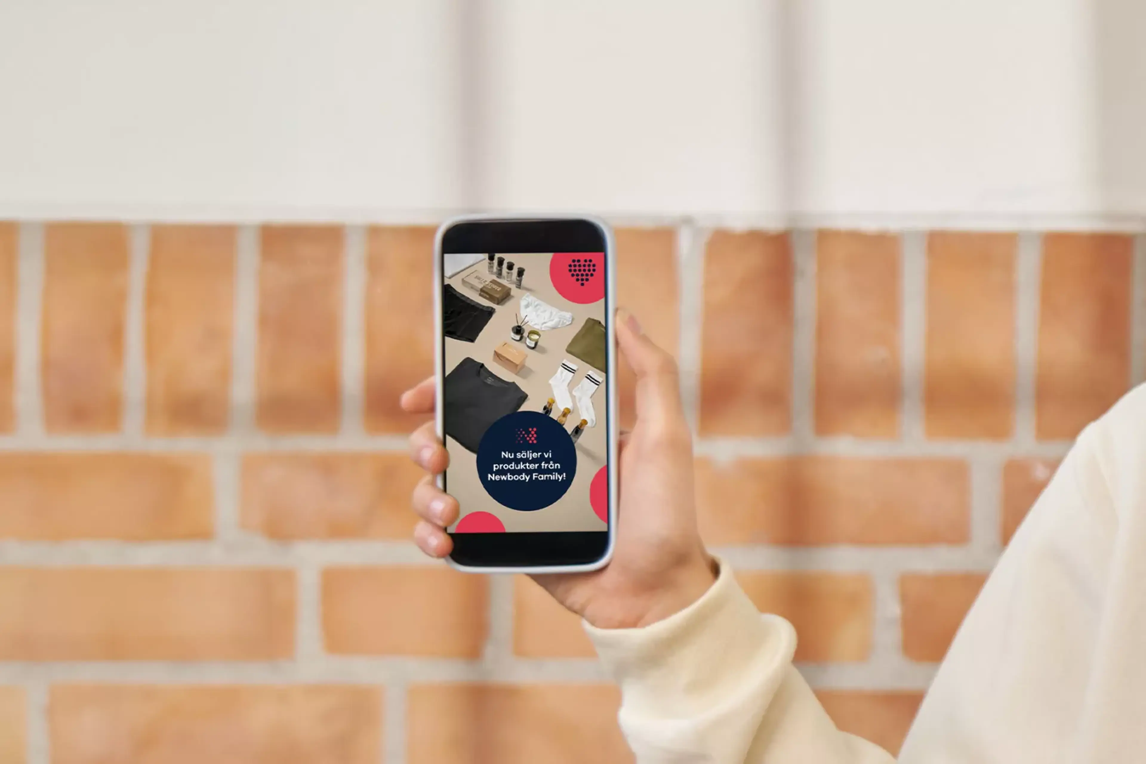 En hand håller upp en smartphone med en digital annons där olika kläder och accessoarer är prydligt uppställda mot en ljus bakgrund. Text på skärmen meddelar att produkter från Newbody Family nu finns till försäljning.