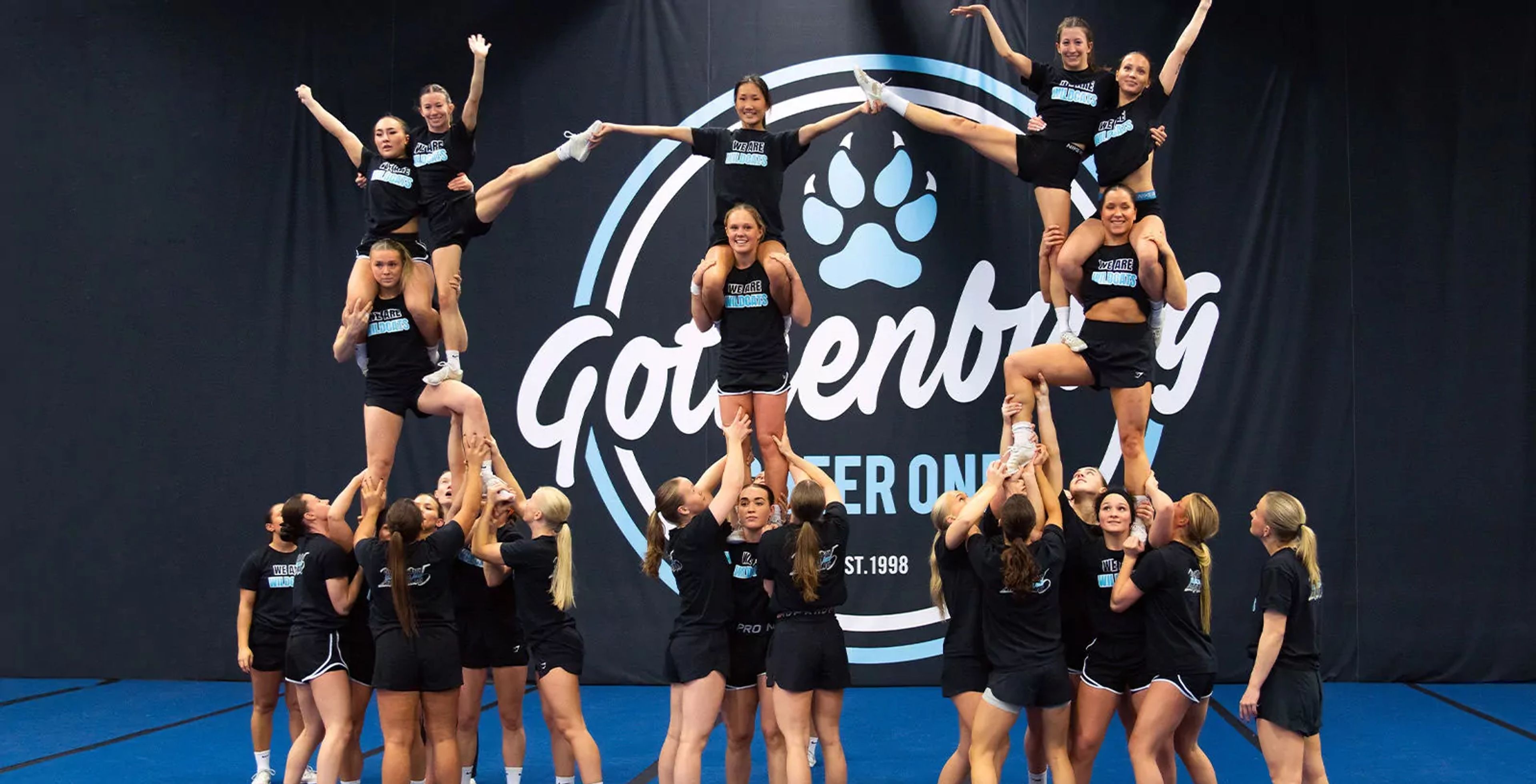 Ett cheerleadinglag i svart träningskläder utför lyft och formationer på en blå matta framför en vägg med texten ”Gothenburg”. Teamet tränar tillsammans med fokus och samarbete.