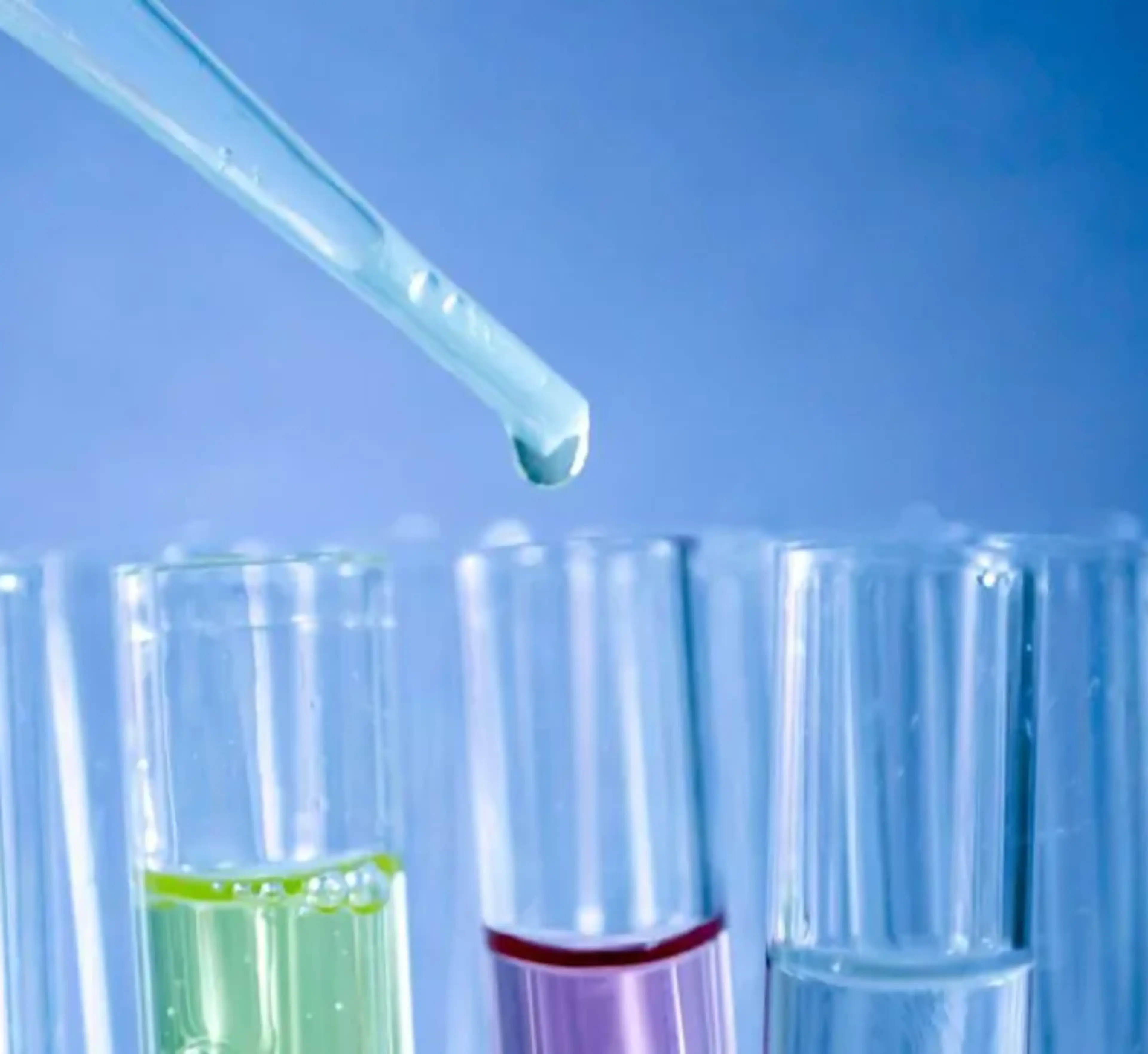 En pipett som droppar vätska över provrör med färgade vätskor i en laboratoriemiljö. En närbild som visar detaljer i glasrören mot en blå bakgrund.