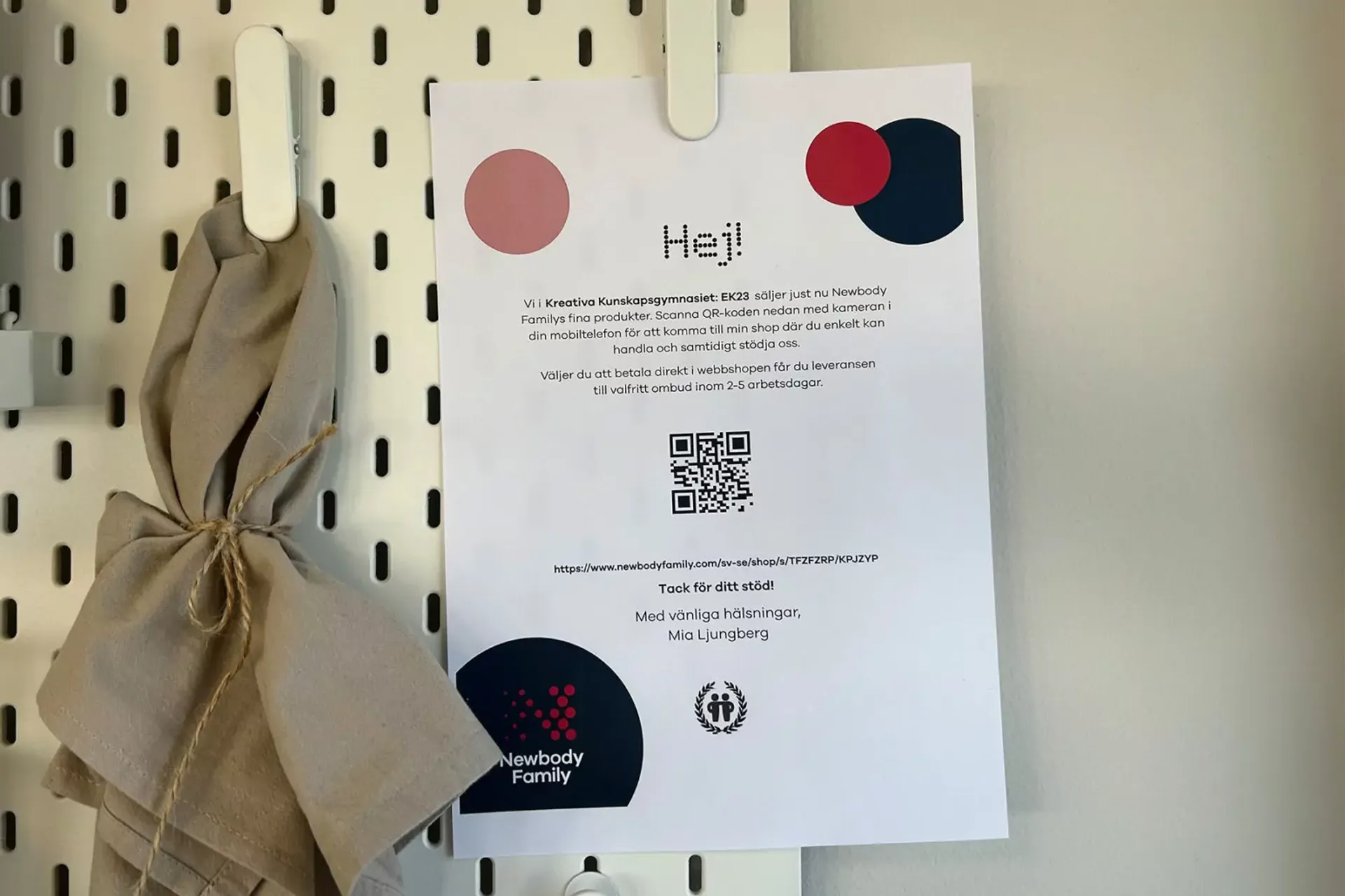 En informationslapp med QR-kod för att handla Newbody Familys produkter online, upphängd på ett vitt perforerat fack med en beige tygpåse bredvid. Texten förklarar support till Kreativa Kunskapsgymnasiet EK23 och hur leverans fungerar