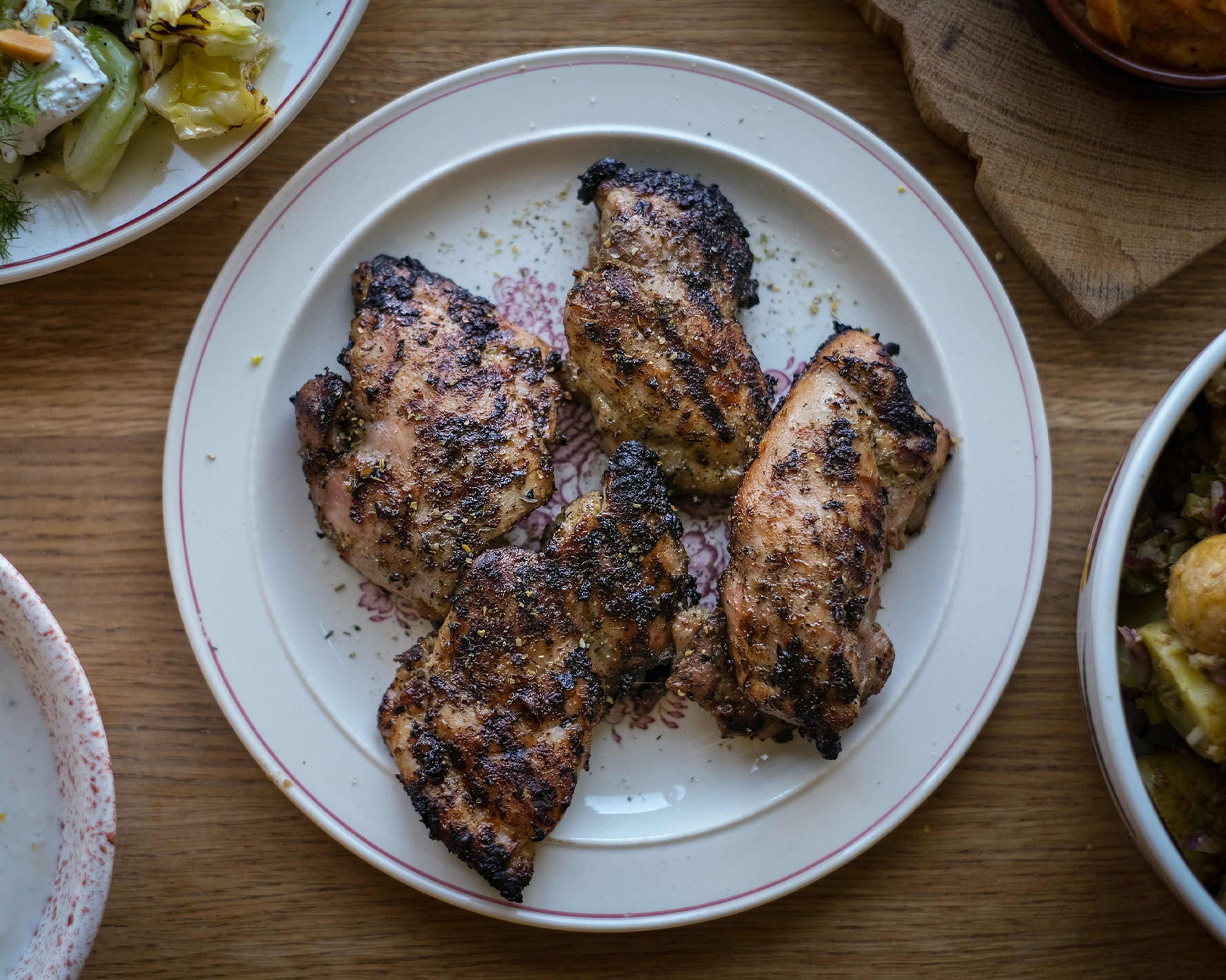 Fyra bitar grillade kycklinglårfiléer med kryddig yta ligger på en vit tallrik med dekorativt mönster. Runtom syns delar av andra maträtter på träbordet.
