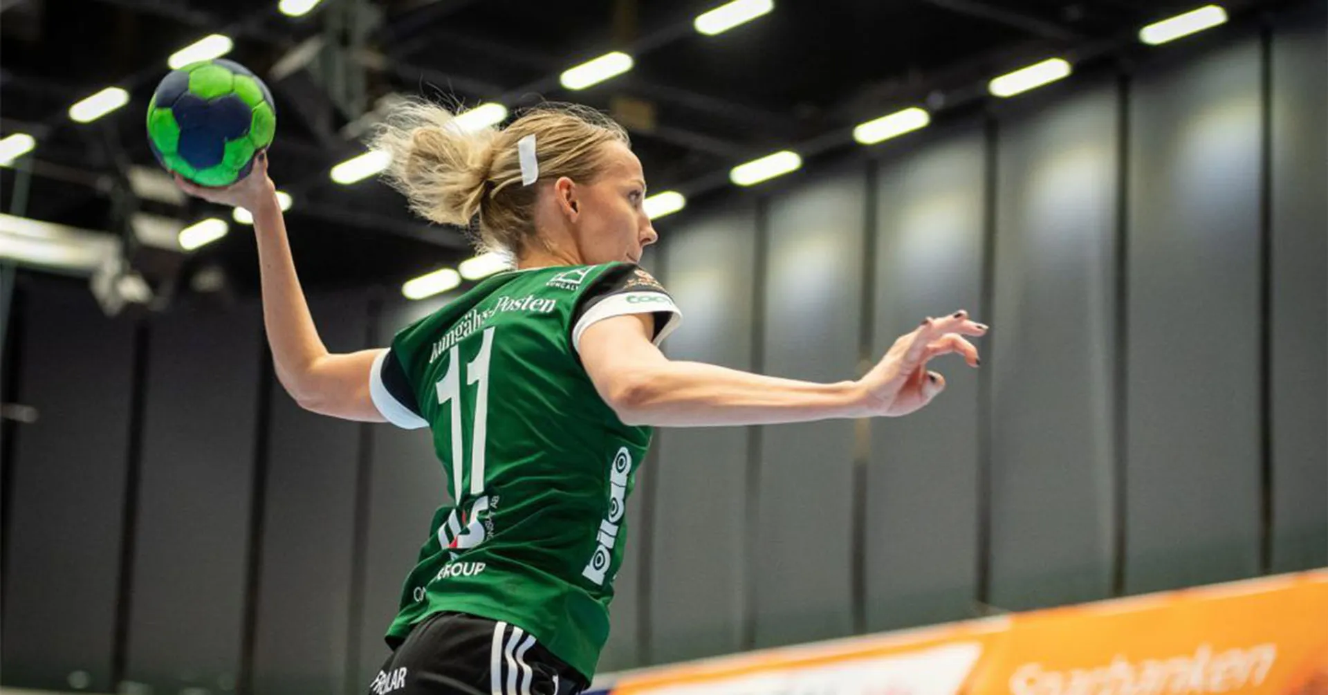 En handbollsspelare i grön tröja hoppar för att kasta en blå-grön boll i en inomhushall. Bakgrunden visar reklamskyltar och högt i tak.