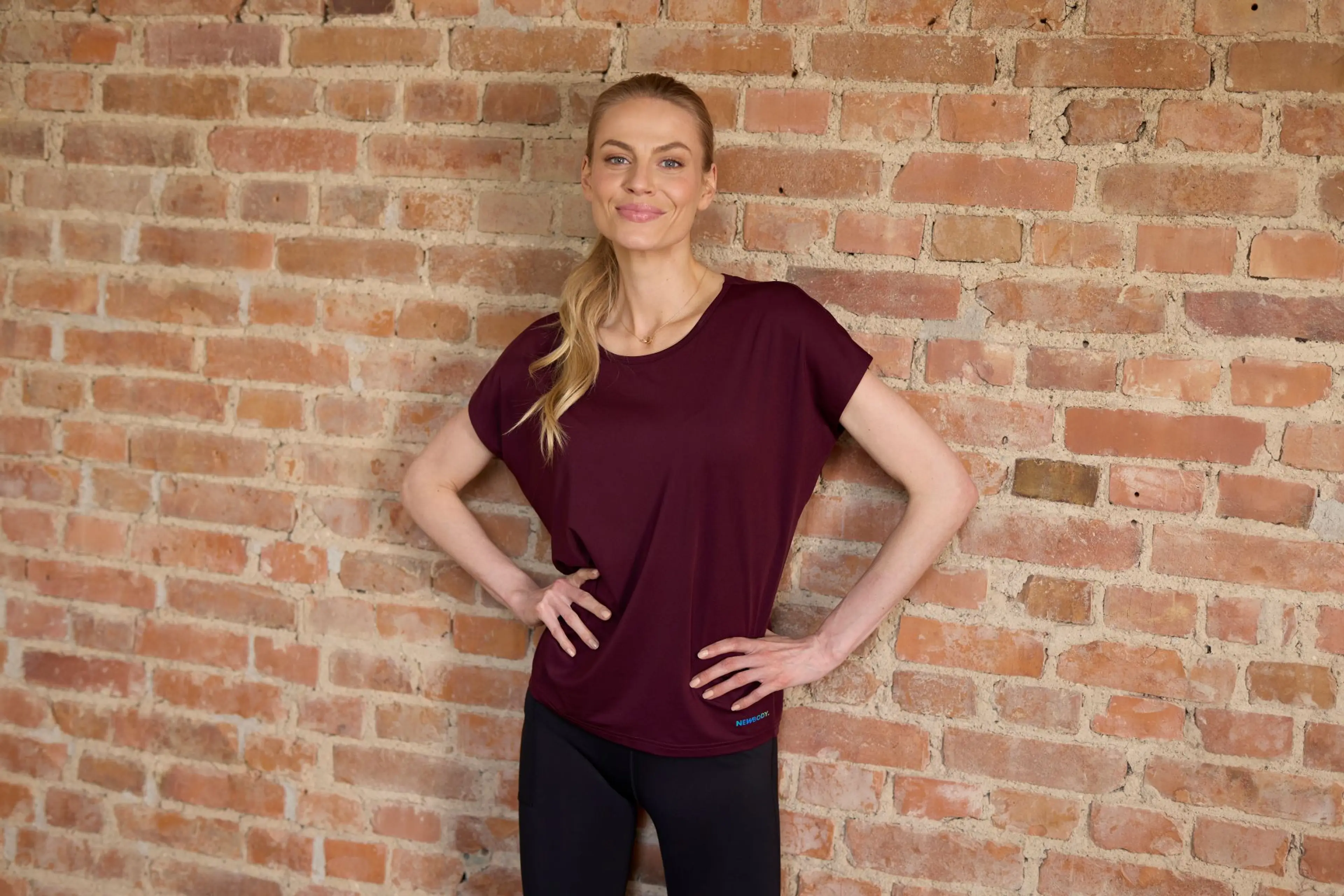 Blond kvinna står mot en tegelvägg med armarna i midjan och bör en vinröd träning t-shirt