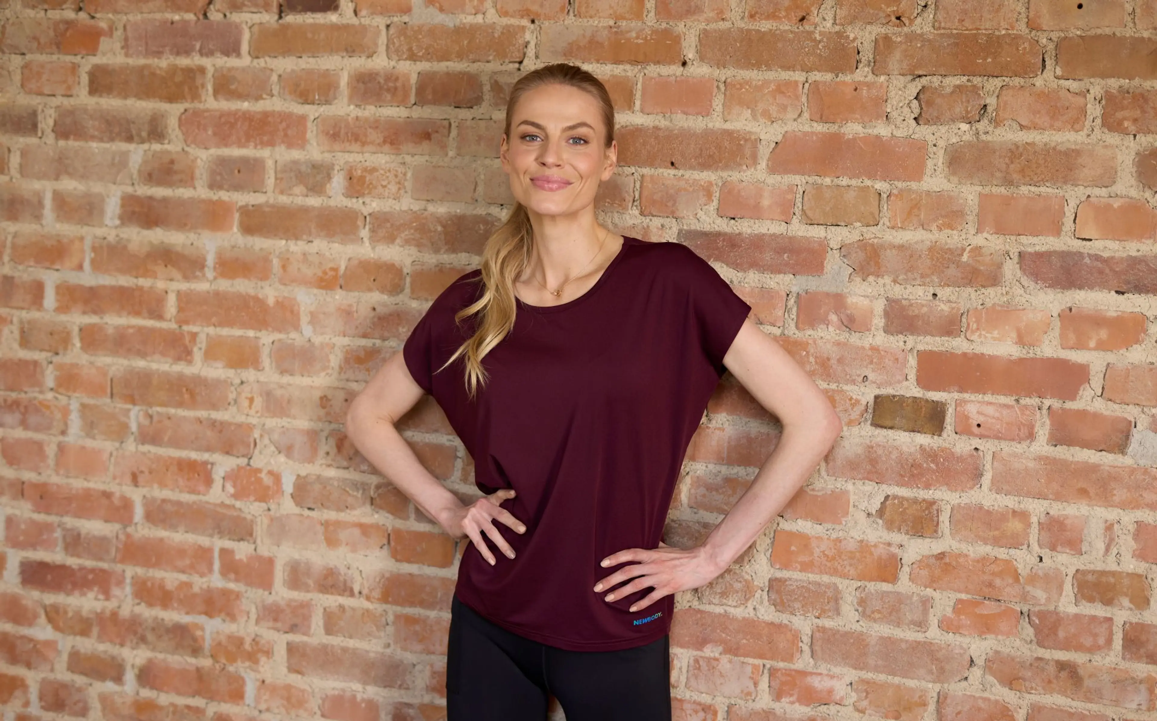 Blond kvinna står mot en tegelvägg med armarna i midjan och bör en vinröd träning t-shirt