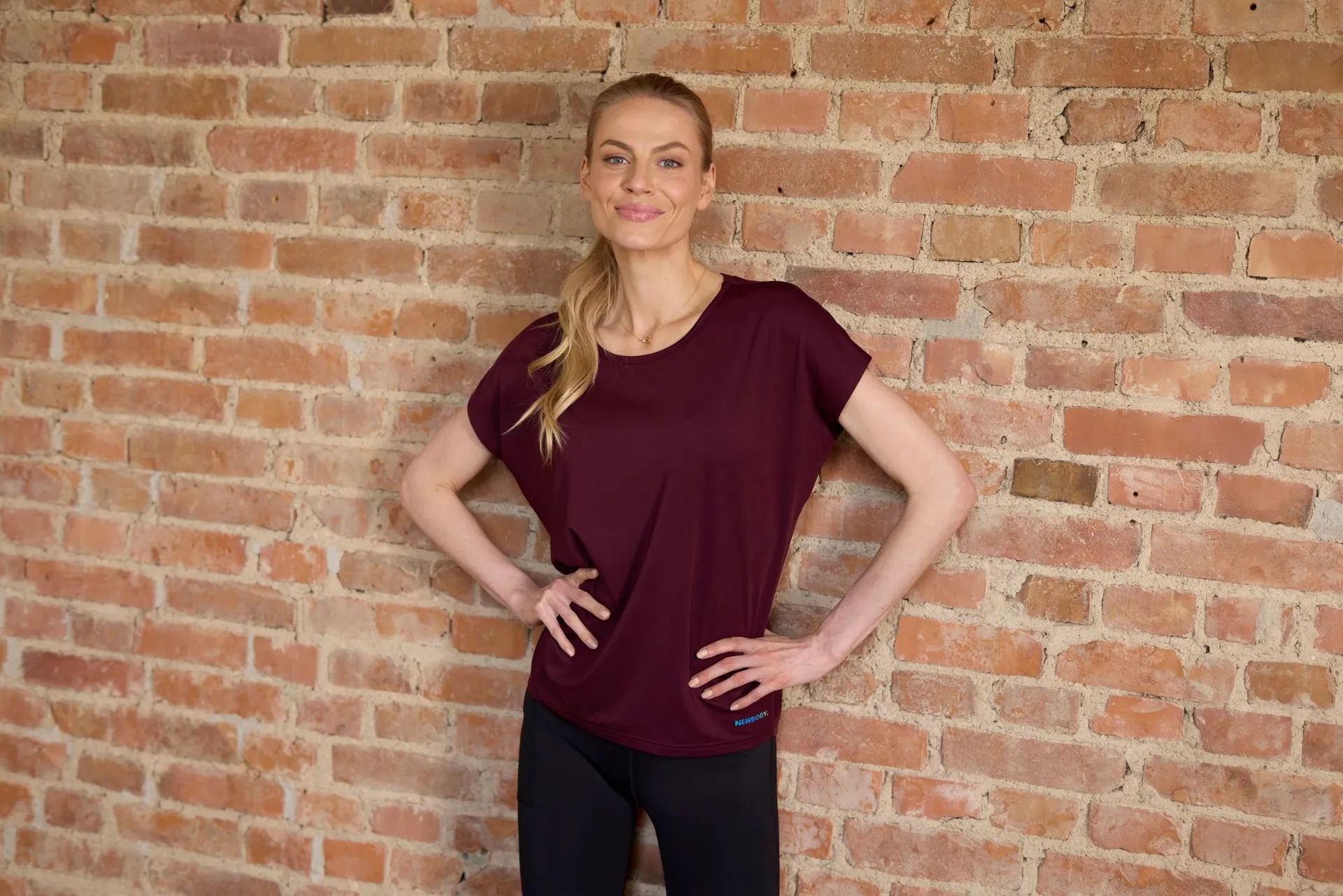 Blond kvinna står mot en tegelvägg med armarna i midjan och bör en vinröd träning t-shirt