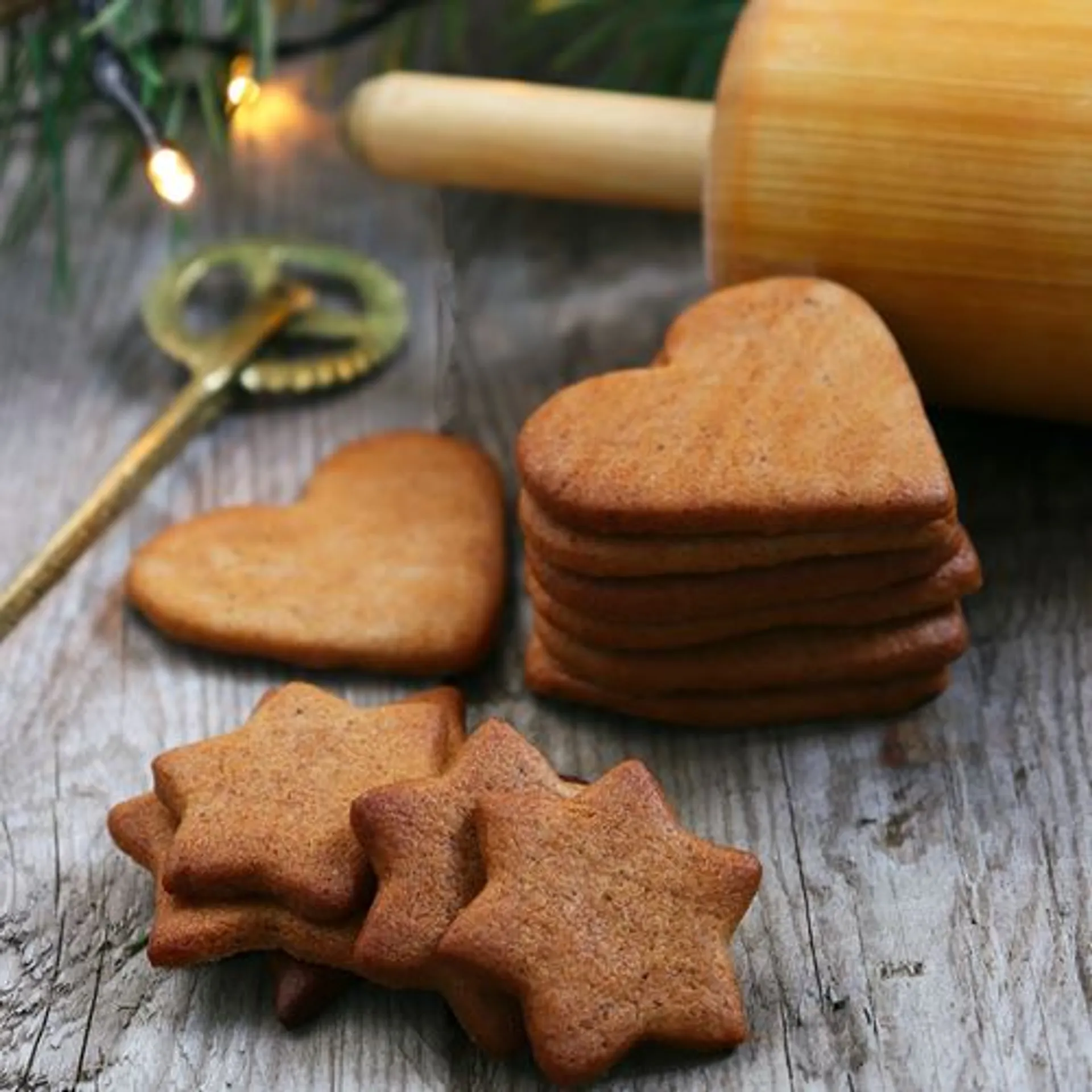 Hjärte- och stjärnformade pepparkakor i en varm brunsvart ton ligger staplade på ett träbord, med en kavel och pepparkaksslev i bakgrunden.