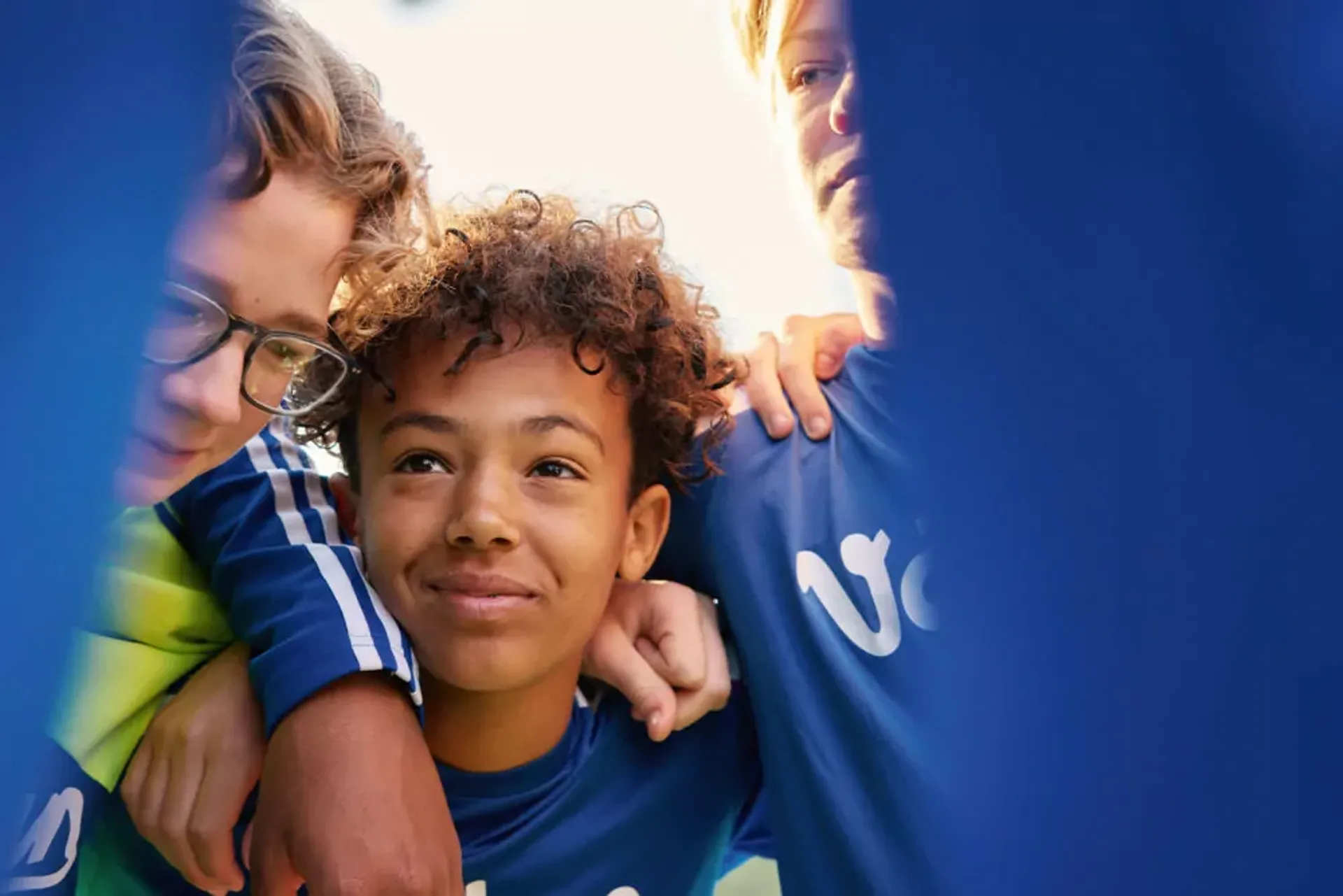 Tre ungdomar i blåa tröjor som står tätt tillsammans, ena personen har armen om en annan. De ser ut att dela en stund av samhörighet och stöd.