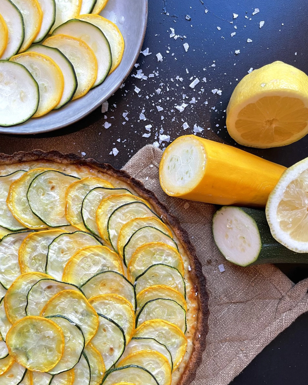 En paj dekorerad med tunna skivor av gul och grön zucchini, med grovt havssalt strött runtom. Skivade citroner och zucchini ligger bredvid på en mörk yta med en beige duk under.