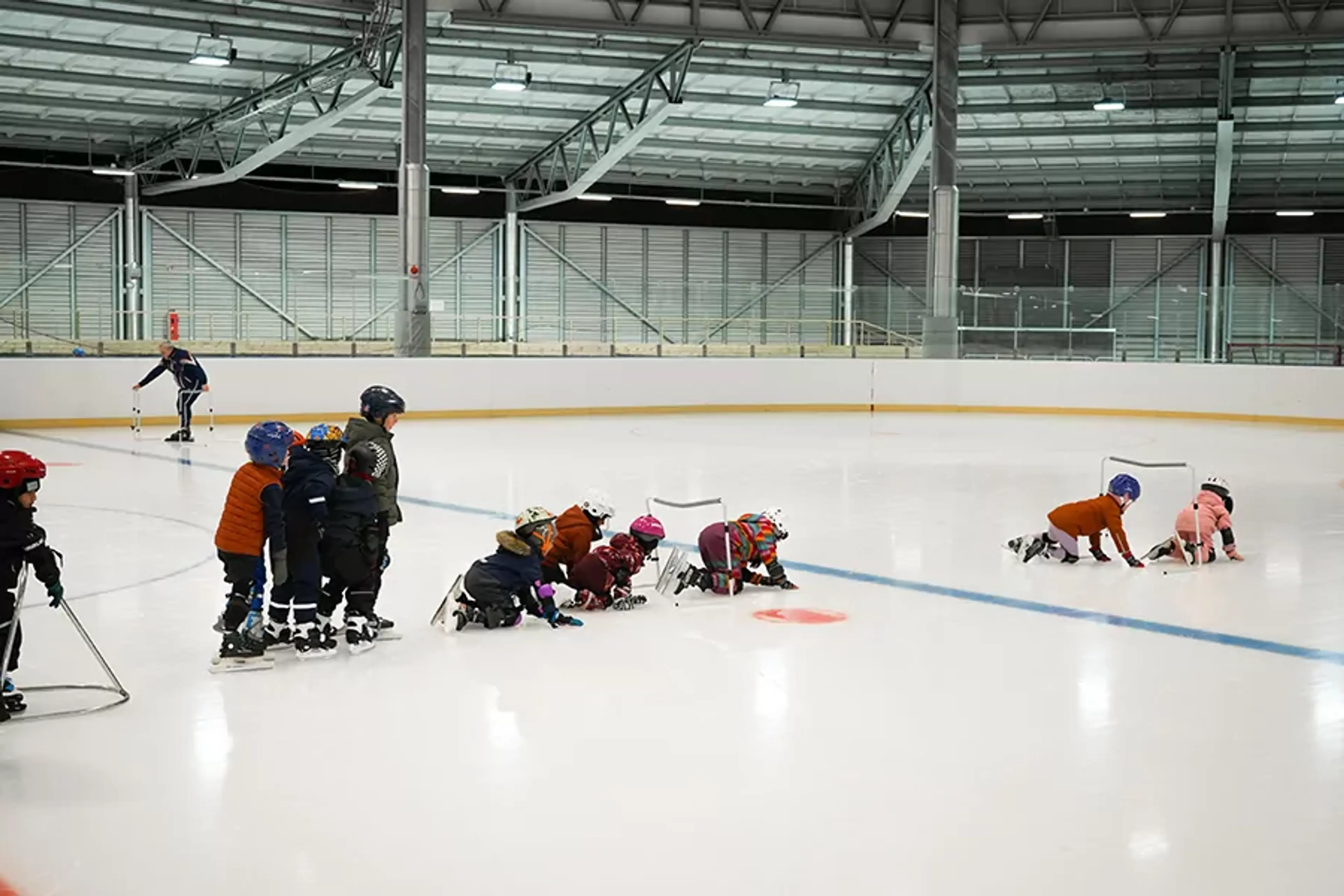 Barn i hjälmar och vinterkläder övar på att åka skridskor på en inomhusrink med hjälp av stöd och handledning. En ledare syns i bakgrunden som följer aktiviteten.
